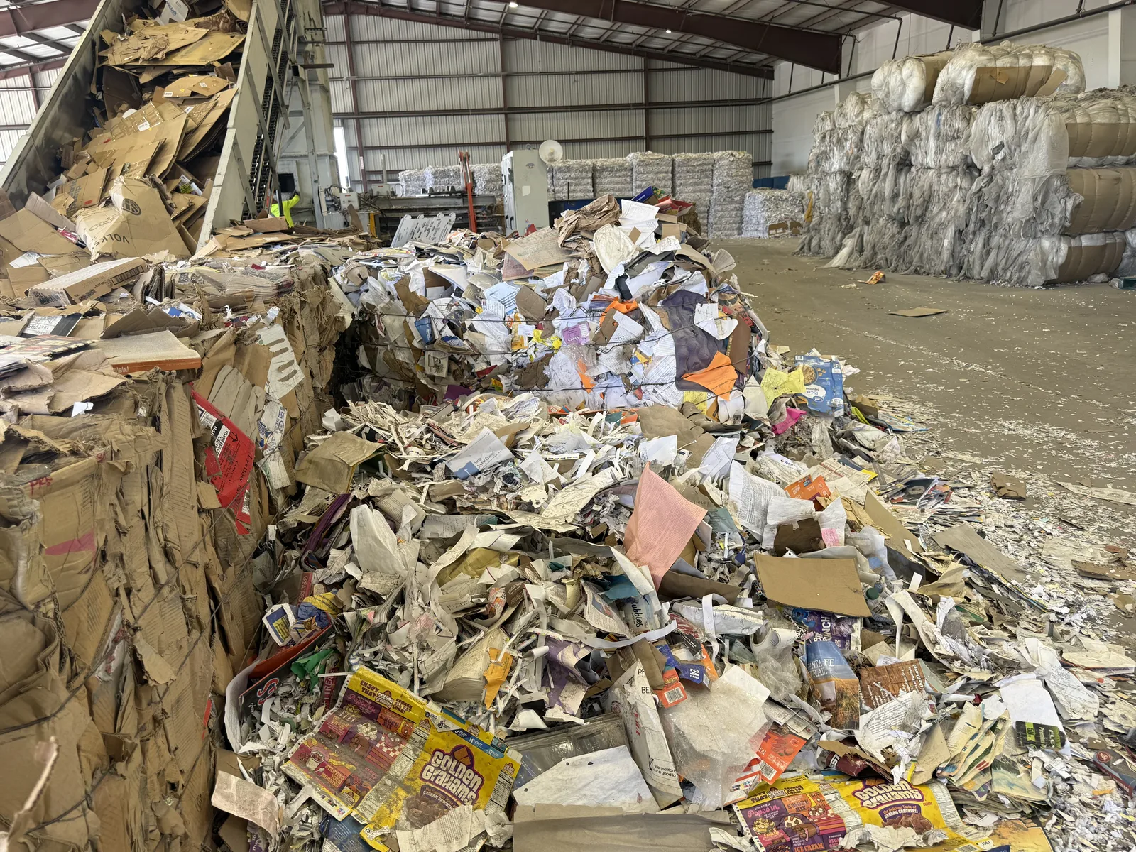 Interior of an Albuquerque paper recycling facility with a mountain of loose books, magazines and mixed paper on the tipping floor beside baled stacks