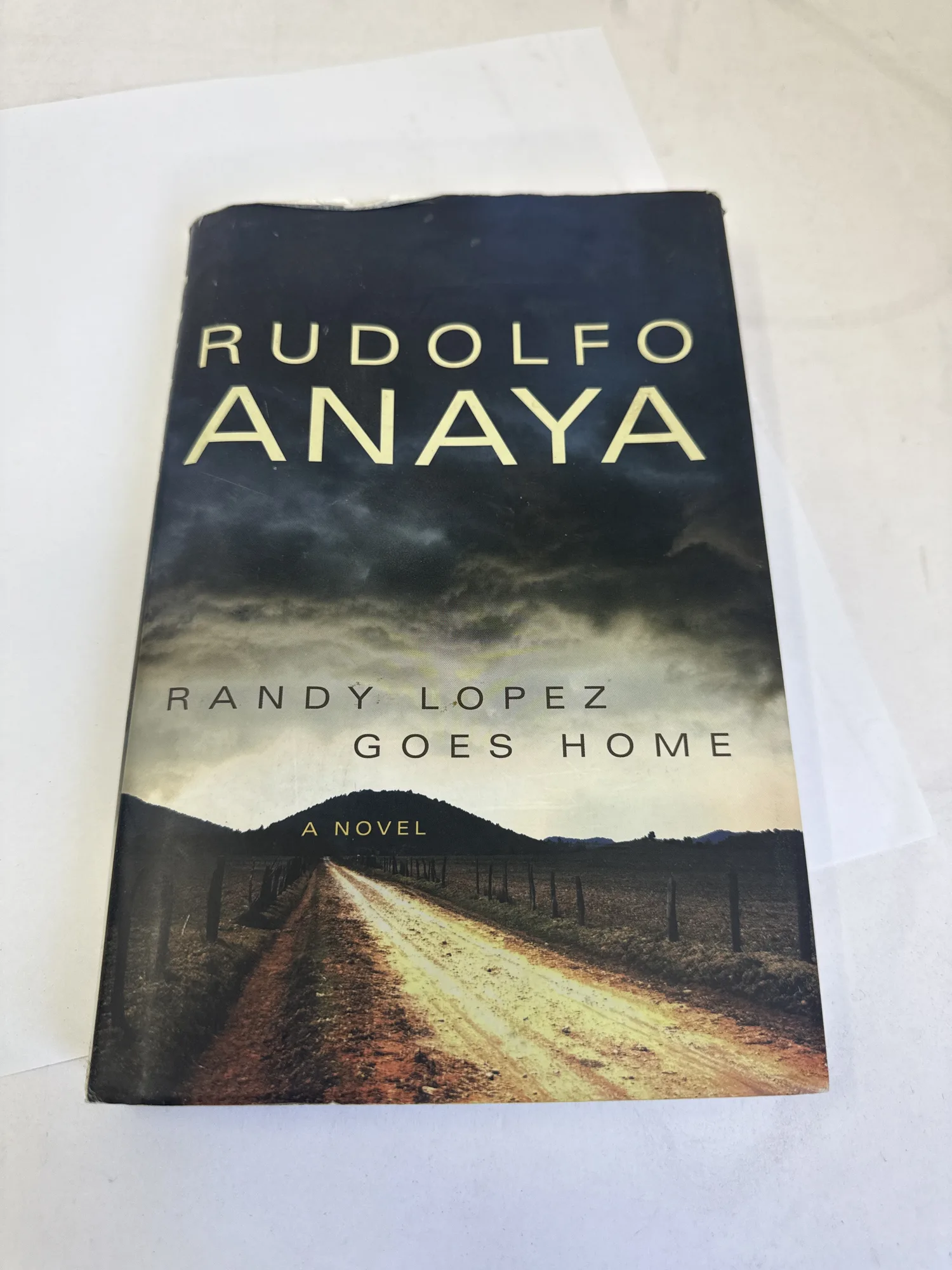 Front cover of Randy Lopez Goes Home by Rudolfo Anaya, first edition hardcover published by University of Oklahoma Press 2011, showing dramatic New Mexico landscape with storm clouds over desert terrain and an adobe structure, author name RUDOLFO ANAYA in large gold serif type. Photographed at the New Mexico Literacy Project sorting desk in Albuquerque.