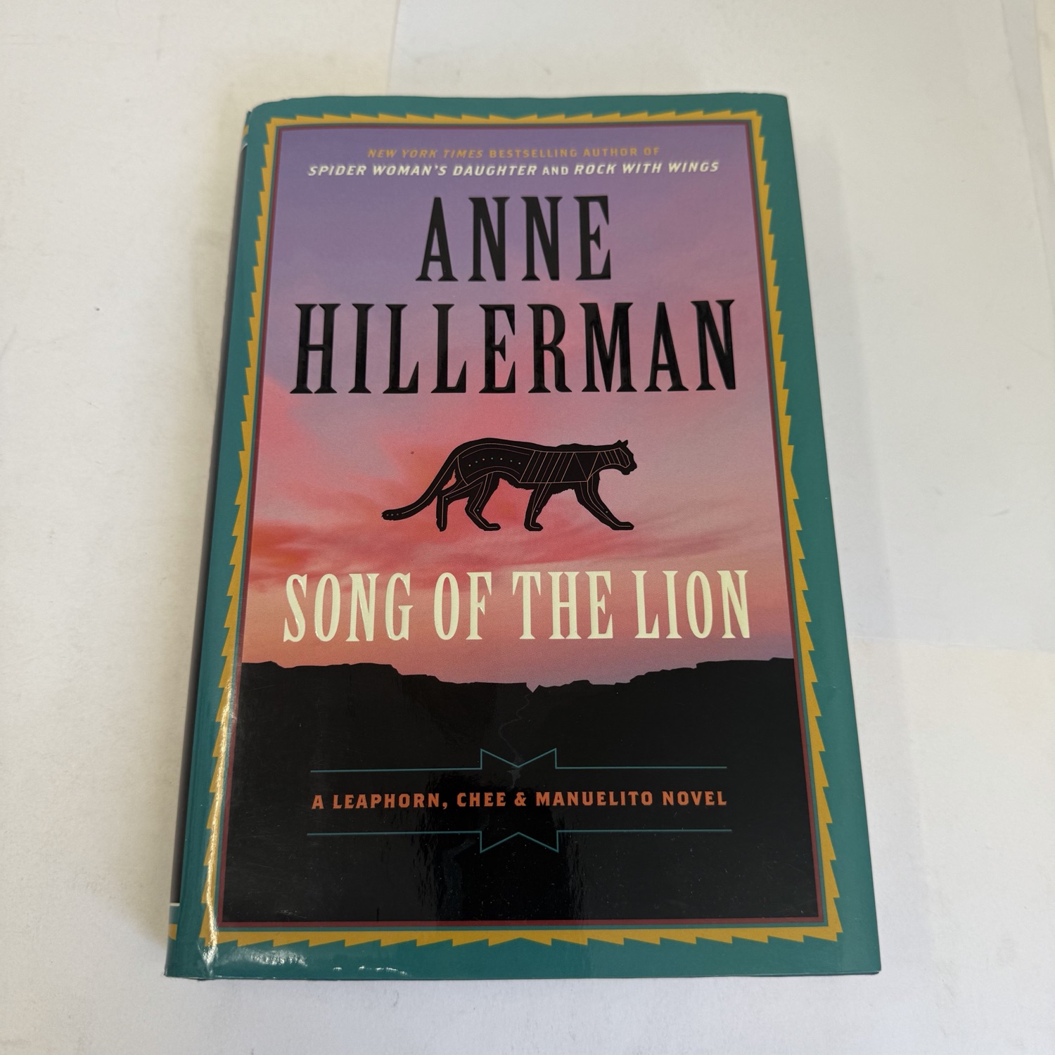 Front cover of Anne Hillerman's Song of the Lion first edition hardcover with dust jacket — turquoise border with Navajo-inspired zig-zag pattern, sunset sky over mesa silhouette, mountain lion silhouette, published by Harper 2017