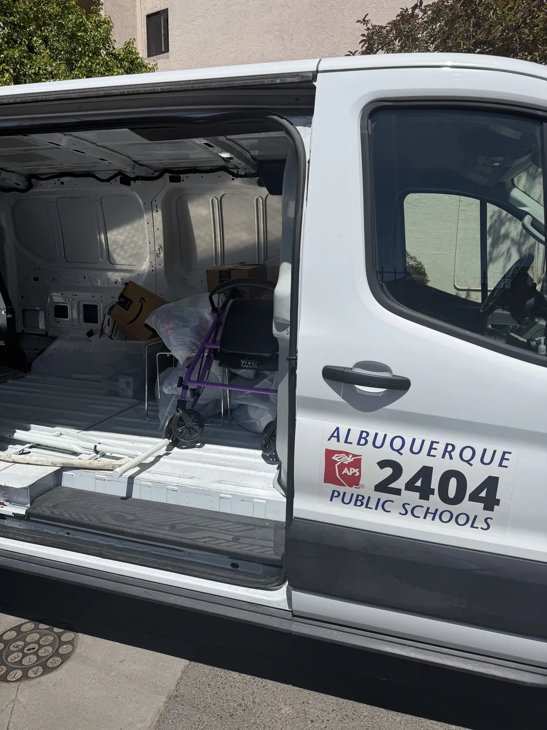Albuquerque Public Schools van #2404 with the APS logo on the door, parked at the New Mexico Literacy Project warehouse with rear cargo doors open and a load of books and supplies inside, secured with purple cargo straps. Photographed during a Tuesday Title I / McKinney-Vento delivery.