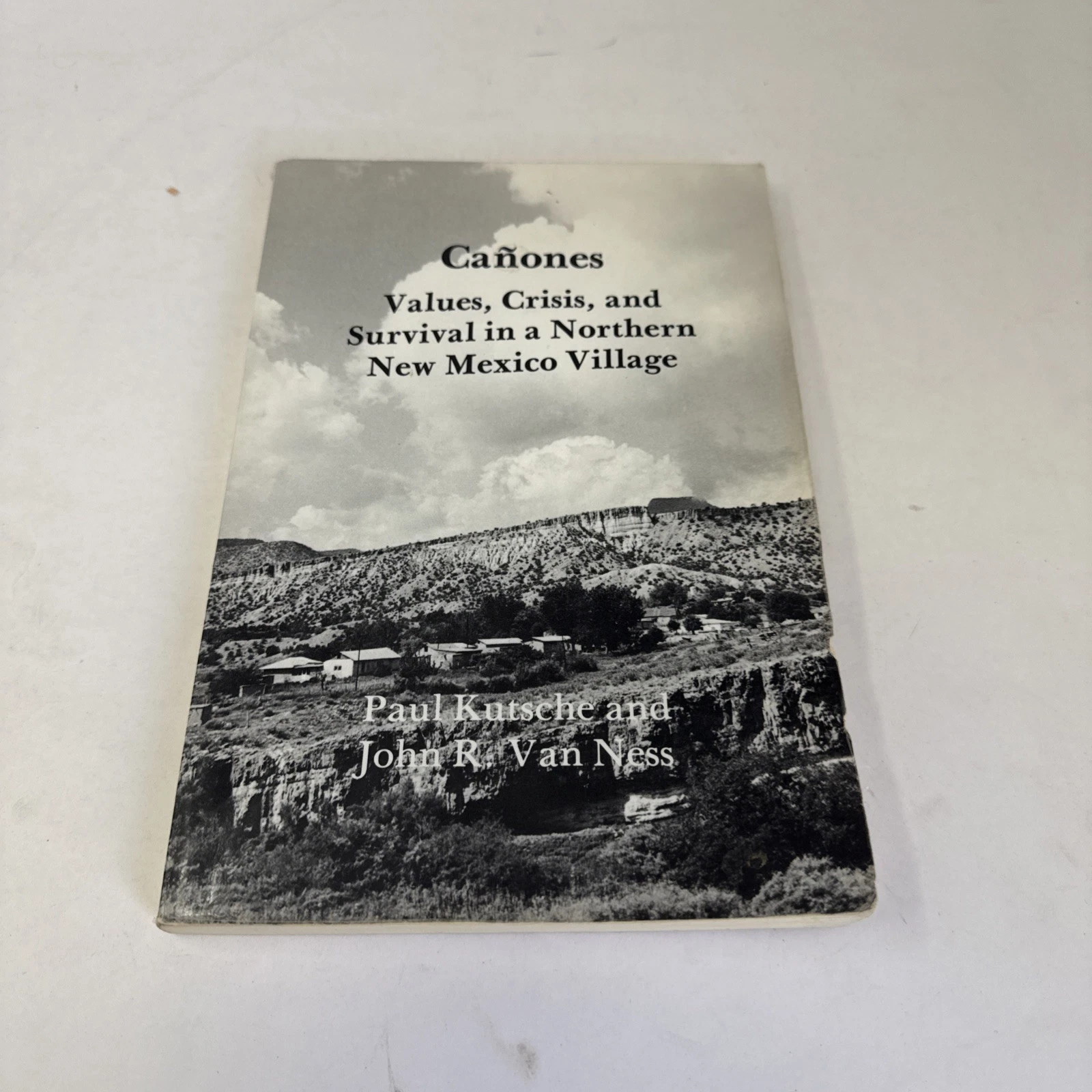 The front cover of the Sheffield Publishing 1988 paperback reissue of Cañones: Values, Crisis, and Survival in a Northern New Mexico Village by Paul Kutsche and John R. Van Ness. Cover layout is plain academic-press paperback: title in serif type at top, authors below, with a black-and-white photograph of the village of Cañones taking up the central two-thirds of the cover — adobe houses and outbuildings clustered in the high-desert landscape of Río Arriba County, with juniper and piñon hills behind.