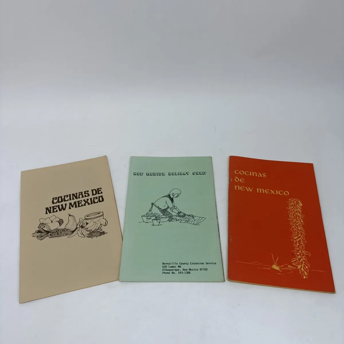 Three saddle-stitched soft-cover regional New Mexico cookbooks lying side-by-side: a tan Cocinas de New Mexico cover with vegetables-and-corn illustration, a green Bernalillo County Extension Service New Mexico Holiday Show cookbook, and an orange Cocinas de New Mexico variant with chile-ristra illustration.