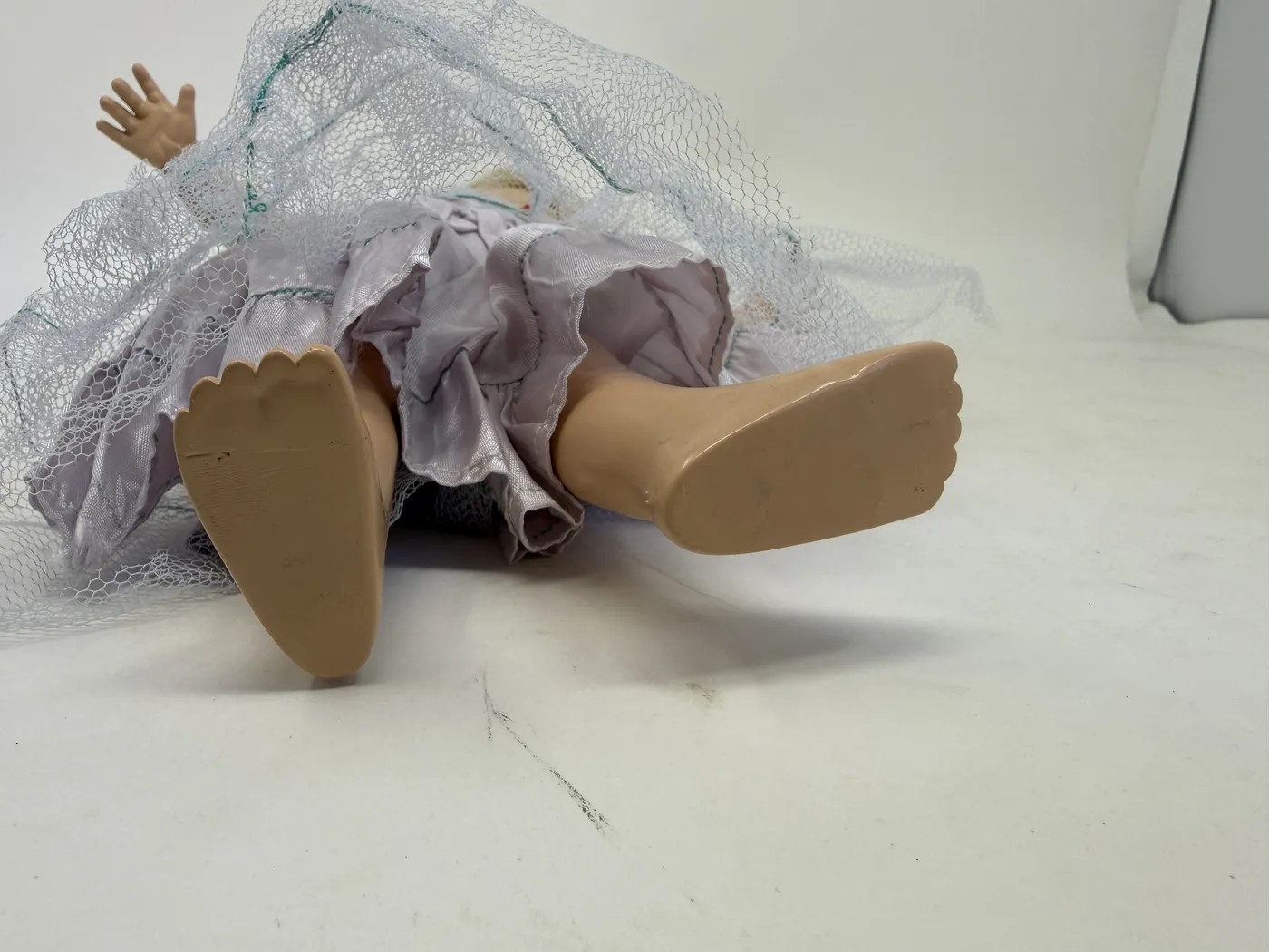 A view of the lower body of the Toni doll with the lavender taffeta dress pulled up to expose the hard-plastic legs and abdomen. The flesh-toned hard-plastic surface is intact and smooth, with the molded-in belly button visible at the abdomen and the molded outline of the hip joint visible at the upper thigh. The legs are jointed at the hip and the seam between the upper body and the leg is visible as a faint horizontal line. The feet are visible at the bottom of the frame with the integrated flat soles. The tulle skirt fans out to either side of the doll's lower torso.