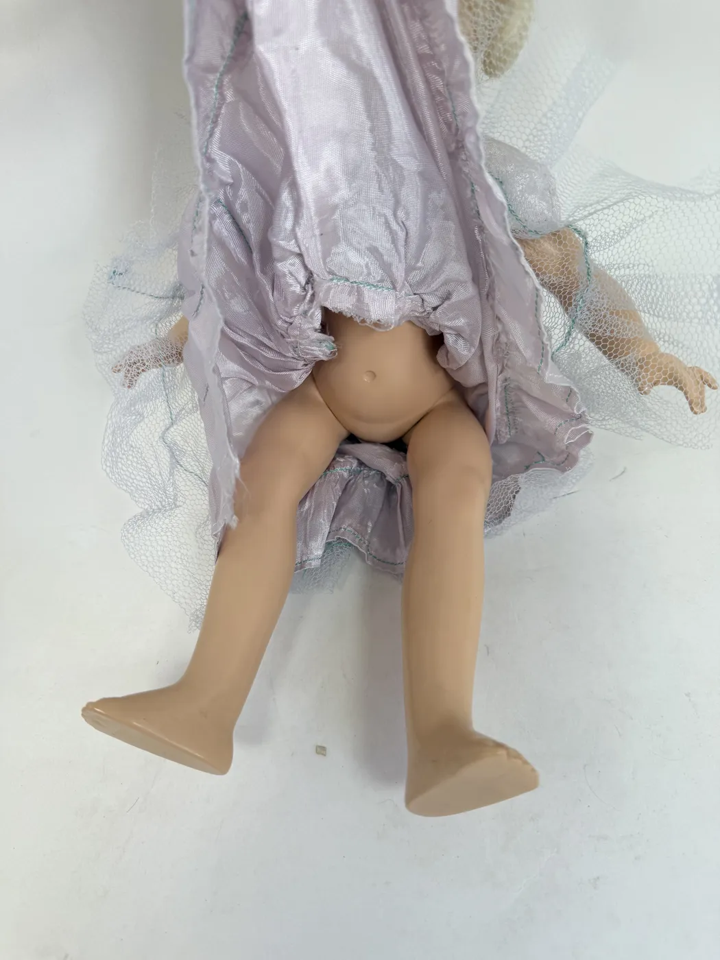 The Toni doll laid on its back, photographed from above, showing the platinum-blonde wig swept up in the back into a softly tucked side roll. The dress is visible from behind: pale lavender taffeta bodice with the sailor-collar back, full layered tulle skirt with teal stitched rows fanning out below. Both arms of the doll are extended out from the body. The overall styling of the wig and dress reads as the late-1940s mass-market doll-fashion aesthetic.