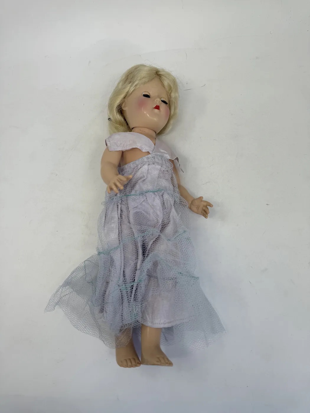 A view from above of the Toni doll lying on its back with the dress pulled fully down off the upper body and exposing the hard-plastic torso. The skin tone is uniform pale flesh-pink across the chest, abdomen, and upper legs. The molded breast and shoulder anatomy is subtle but visible. The arms are jointed at the shoulder and visible folded against the sides of the torso. The platinum-blonde wig is visible above the head, slightly disarrayed from the undressing.