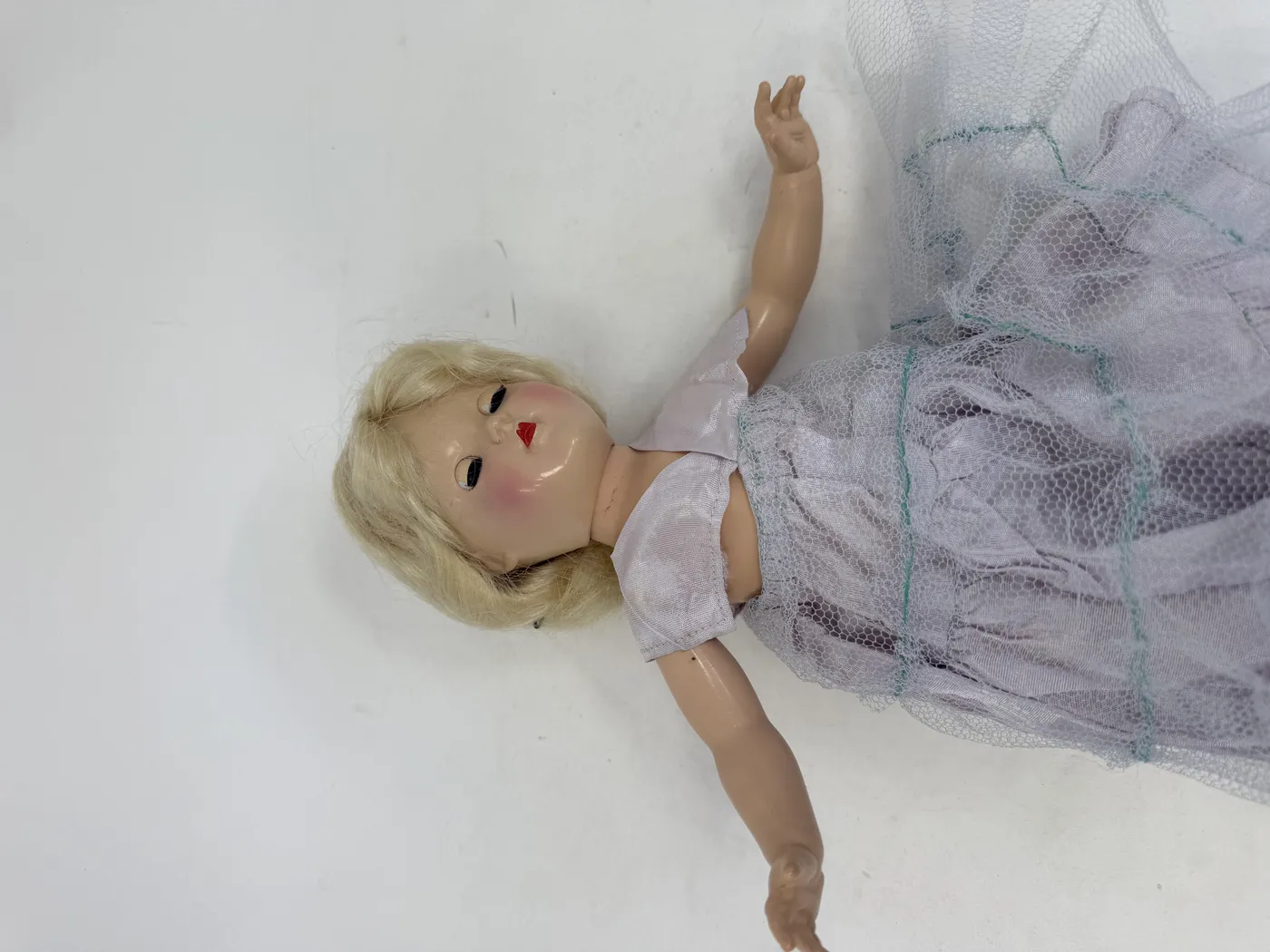 The Toni doll lying on its back, photographed from above. The head is tilted slightly to the left with the platinum-blonde wig spreading on either side of the head, the lips parted slightly open as the doll's mouth-painting is visible from above. The lavender taffeta bodice with V-neck sailor collar is visible at the upper torso, the layered tulle skirt fans out around the lower body, and one arm extends out to the side. The hard-plastic flesh-tone is uniform across the visible torso, arms, and legs.