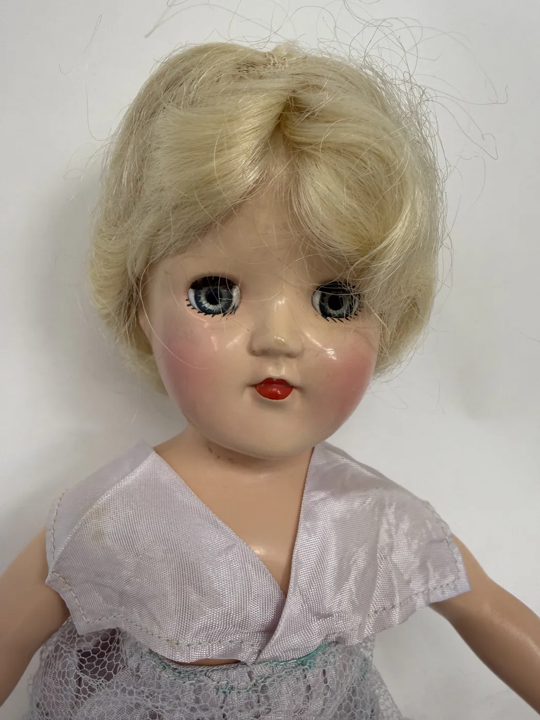 A close-up portrait of the Toni doll's head photographed from the front. The face is hard plastic in a smooth pale flesh-pink finish. Features include large round blue sleep eyes with finely painted upper and lower lashes radiating from each eye, soft pink-blush cheeks blended in oval circles, a small pertly modeled nose, and a small bright red painted bow-mouth set in a closed-lip expression. The wig is platinum blonde in side-parted short-bob nylon fiber, slightly tousled, the hairline meeting the forehead at a natural curve. The pale lavender taffeta V-neck sailor-collar of the dress is visible at the bottom edge of the frame.