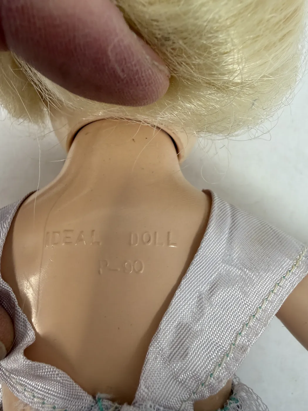 A close-up of the Toni doll's head with the sleep eyes closed, photographed with the doll laid on its back. The closed eyelids are visible as horizontal slits painted with fine black lash detail; the underlayer of painted lower-lash detail shows in soft greyish black at the lower lid line. The cheeks are flushed soft pink. The bow-mouth is painted bright red in a small closed shape with a defined Cupid's bow. The platinum-blonde wig falls naturally on either side of the temples. A faint hairline shadow above one eyebrow indicates a small molded variation in the plastic surface.