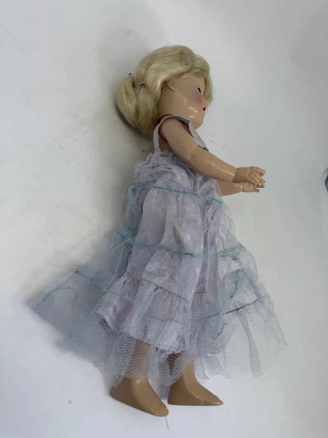 A close-up of the back of the Toni doll's head and the upper back, photographed with the platinum-blonde wig pulled up by a thumb to expose the hard-plastic surface underneath. Two separate molded-in markings are visible, both clearly raised text in the original plastic. The upper marking on the back of the head reads, in three lines: P - 90 (top), IDEAL DOLL (middle), MADE IN U.S.A. (bottom). The neck joint with the body is visible below this marking. The lower marking on the upper back of the body, between the shoulder blades, reads, in two lines: IDEAL DOLL (top), P-90 (bottom). The lavender taffeta V-neck of the dress is visible to the left and bottom of the frame.