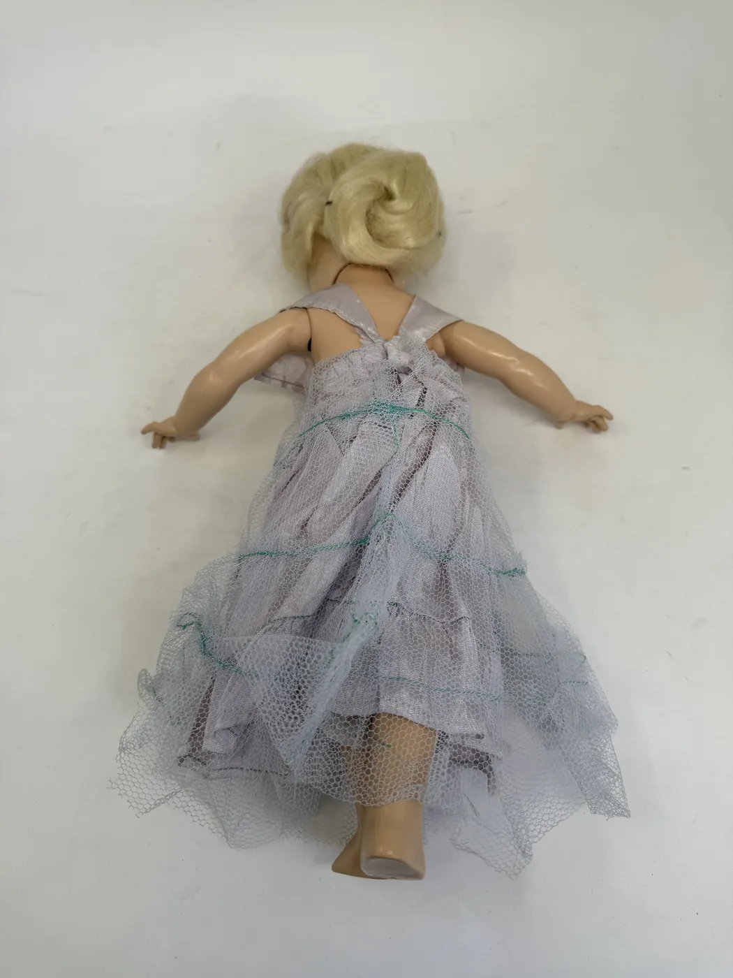A second lying-down view of the Toni doll from the left side. One arm of the doll is raised in front of the face with the palm visible, casting a small shadow on the cheek. The platinum-blonde wig falls in a soft side-tuck against the white background. The lavender taffeta dress with the layered tulle skirt is visible in side profile. The hard-plastic legs extend out from under the dress at the bottom of the frame.