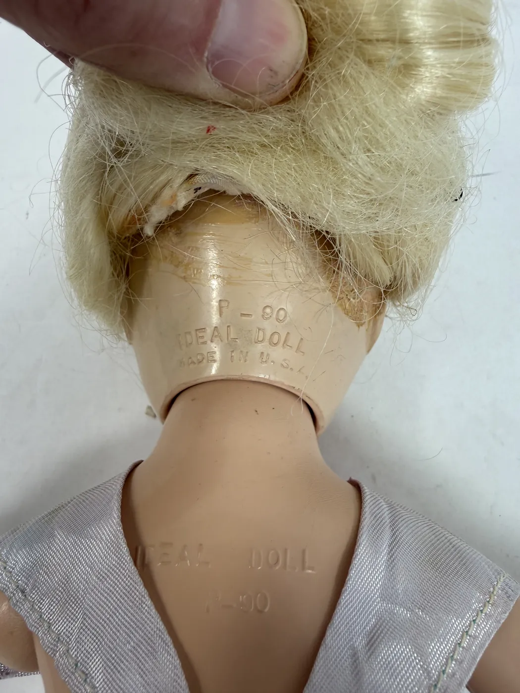 The Toni doll standing in side profile, photographed from the right side. The hard-plastic body is visible from this angle as a continuous flesh-pink color from neck to feet; the doll's right arm hangs at the side with the palm slightly forward, while the head is in profile with the platinum-blonde wig in soft waves over the temple. The lavender taffeta dress with the layered tulle skirt is visible in side profile, falling to mid-calf. The doll is supported on the integrated flat-soled hard-plastic standing feet.