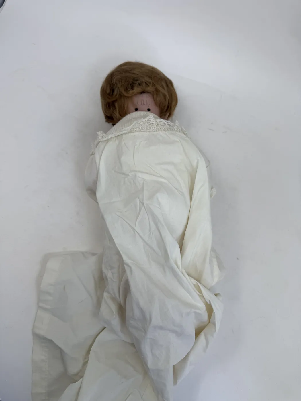 The back of the doll's head and upper body. The mohair wig sits naturally over the bisque, with the side part visible at the crown and the hair sweeping forward and down to a slightly tousled fringe at the nape of the neck. The lace-trimmed collar of the white christening gown is visible below. The doll is laid on its back on a plain white surface. The overall presentation suggests the wig has been on this head for a long time and has not been redressed or recently restyled.
