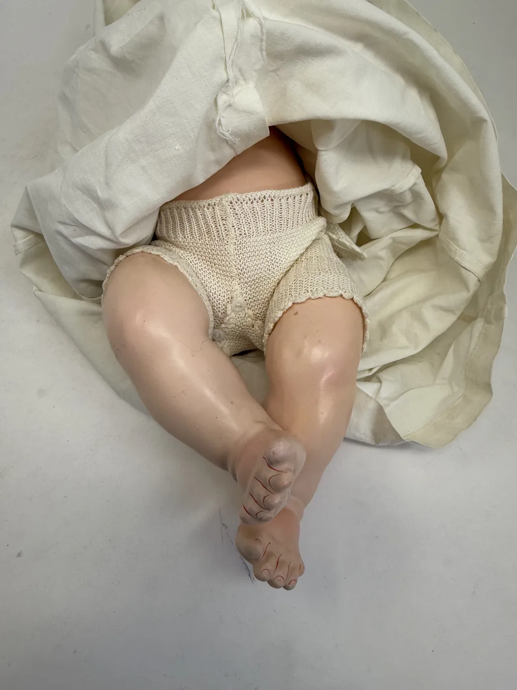 The doll lying on its back with the long christening gown pulled up to expose the lower body. The body is a composition bent-limb baby body in a warm pinkish-flesh paint, modeled with smooth rounded thighs and chubby calves and bent-leg knees, with composition feet showing modeled small toes. The doll is wearing an ivory-cream knit cotton or wool diaper-style romper covering the hips and upper thighs, fastened with two small pearl buttons at the crotch front. A faint factory mold-line is visible running down the center of the upper thigh. The feet show painted-on red detail at the toe-line where the toes meet the foot, a survival of the original paintwork.