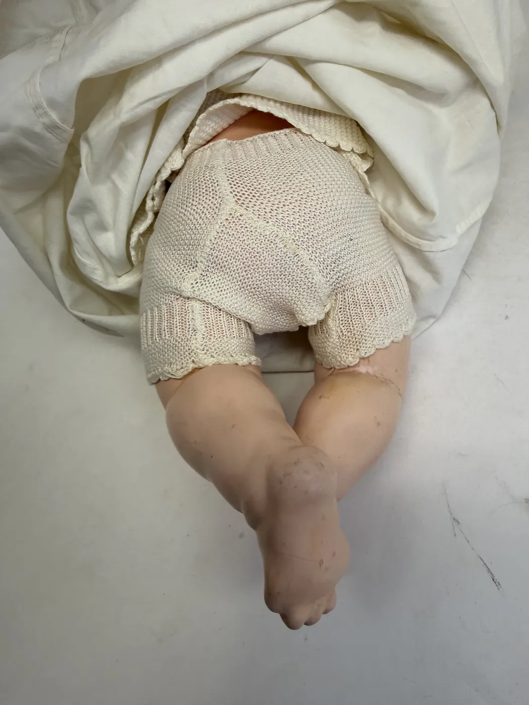A second view of the composition lower body and legs in the knit romper. The composition surface shows the characteristic mild discoloration of hundred-year-old composition: faint craquelure of the paint, some small areas of darker tone where dirt has settled into surface depressions, and the painted toe-line detail surviving on both feet. The mold seam at the inner thigh is visible as a thin pale line.
