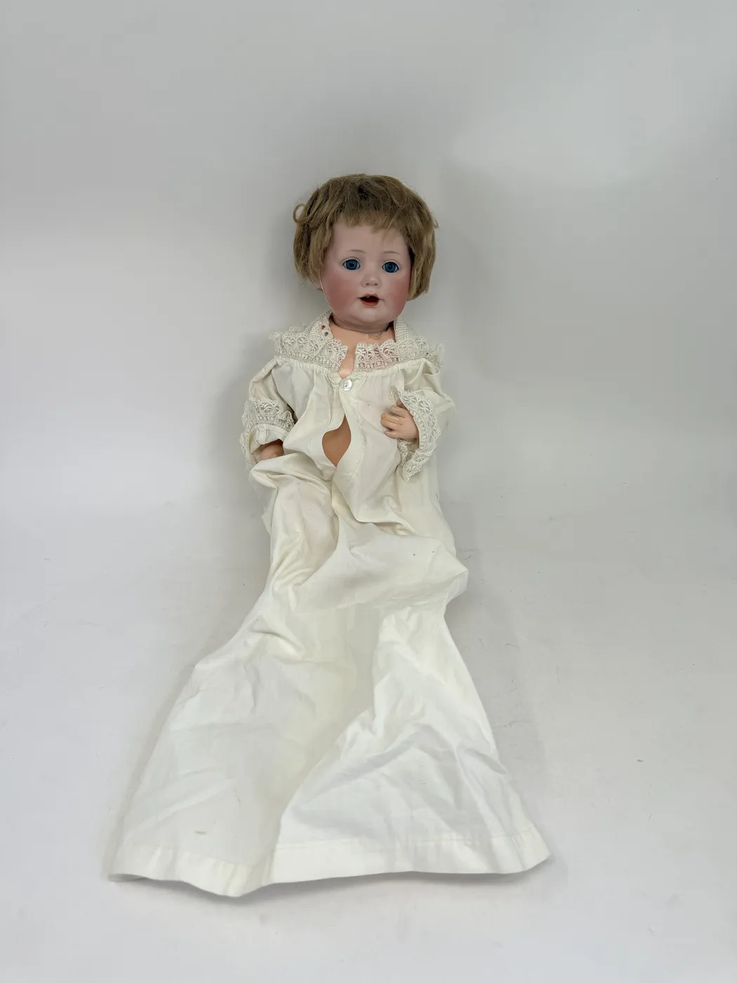A German bisque-headed character baby doll in a long white lace-trimmed christening gown, lying full-length on a plain white surface, with a short brown mohair wig, blue sleep eyes, and an open mouth with two upper teeth
