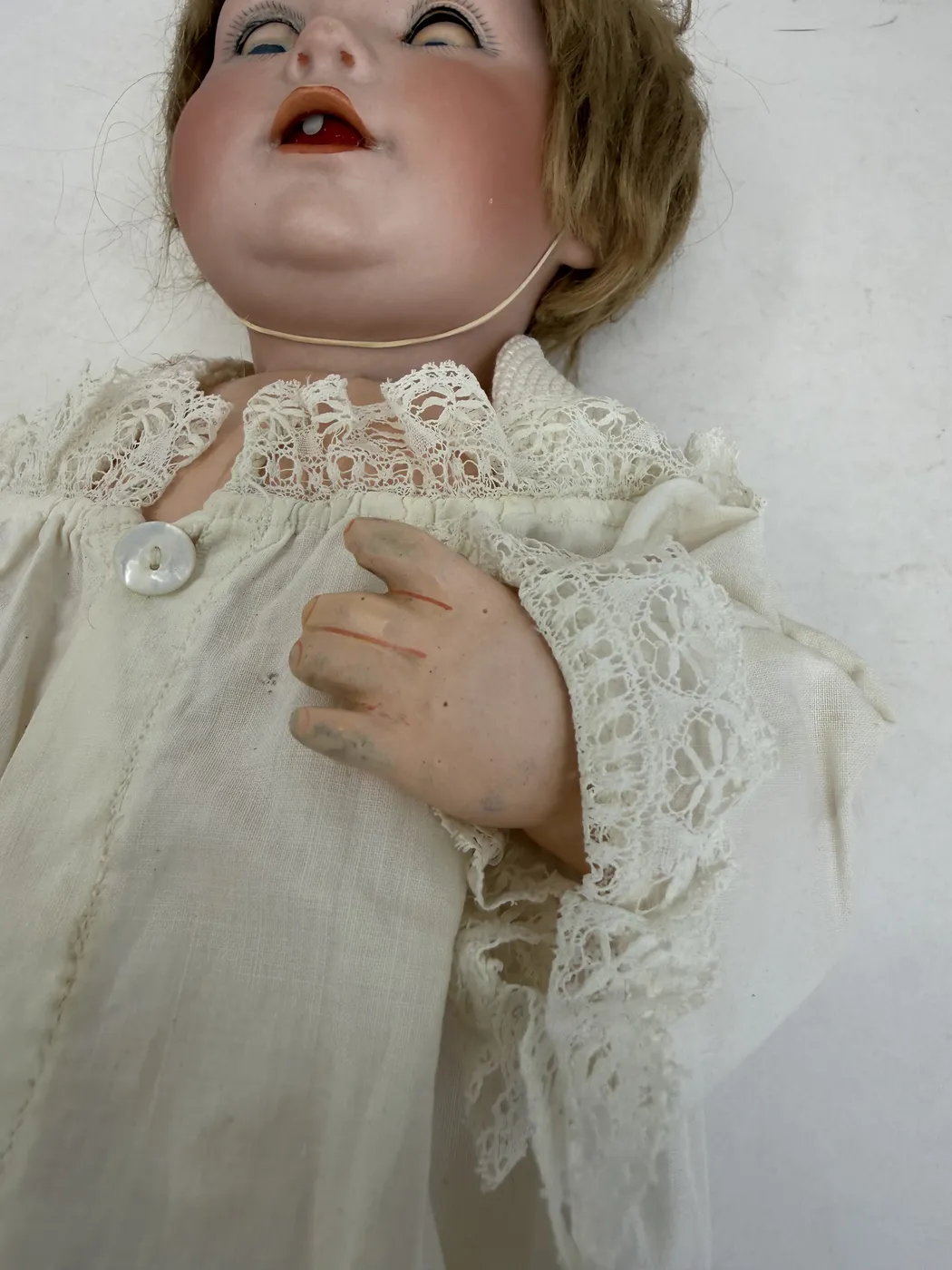A close-up of the doll's right hand resting on its chest above the lace insertion of the christening gown. The hand is composition in a warm flesh tone with each finger separately modeled. Painted-on detail is clearly visible: fine red lines at the joints of each finger, and pronounced red lines at the base of each fingernail. Some of the fingertips show darker oxidation against the original paint, a survival of original paintwork rather than later restoration.