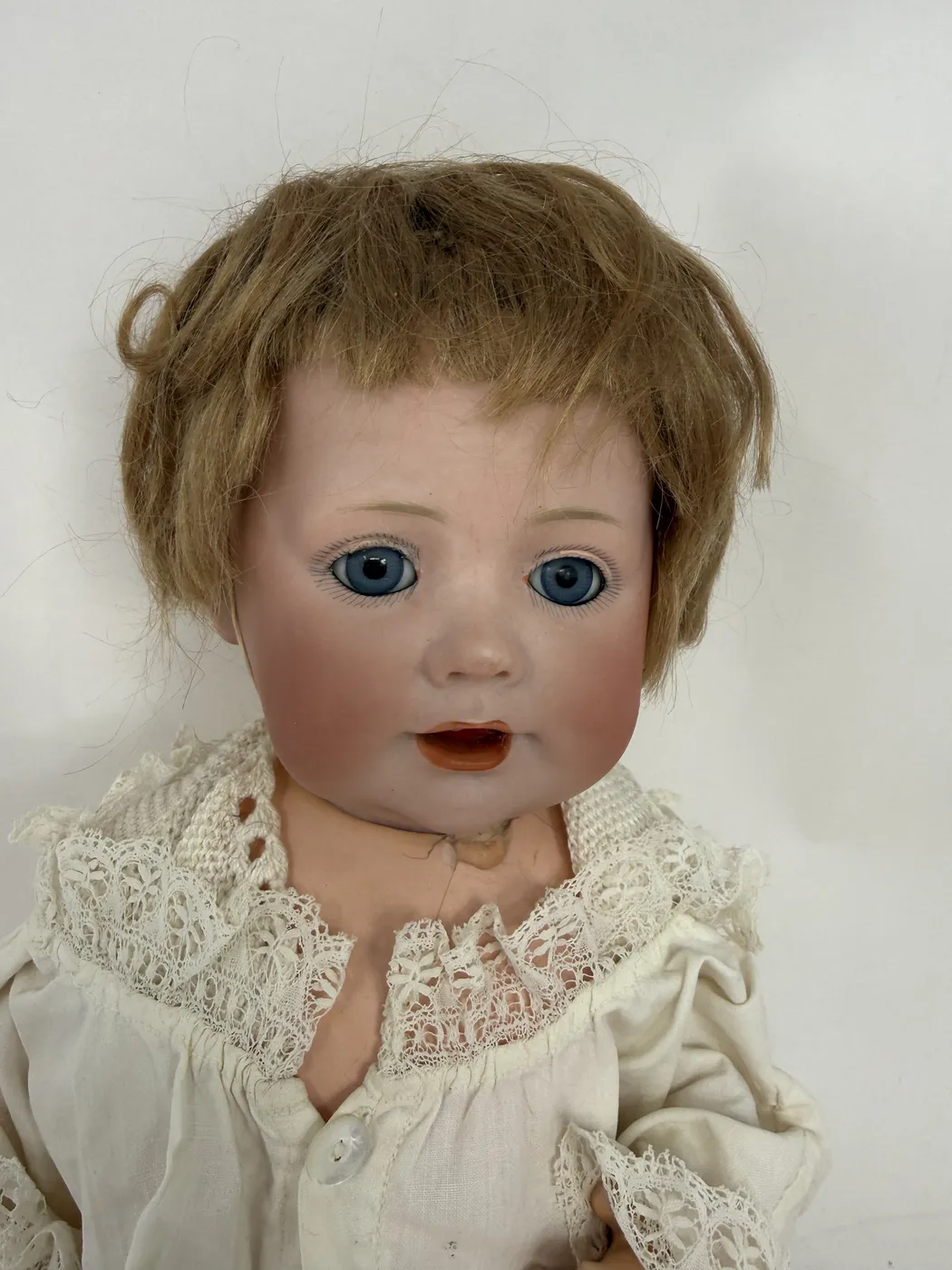 A close-up portrait of the doll's head photographed from the front. The doll's face is rounded with chubby modeled cheeks, a small upturned nose, deep blue sleep eyes with finely painted lashes radiating around each eye, faint single-stroke brows in light brown, and an open mouth showing two small molded upper teeth with a slightly visible orange-pink tongue. The lips are colored a deep matte coral. The mohair wig is light brown and slightly tousled, swept across the forehead in a center-to-side part. A scalloped white cotton-and-lace collar of the christening gown is visible at the bottom of the frame and a single mother-of-pearl button closes the gown at the chest.