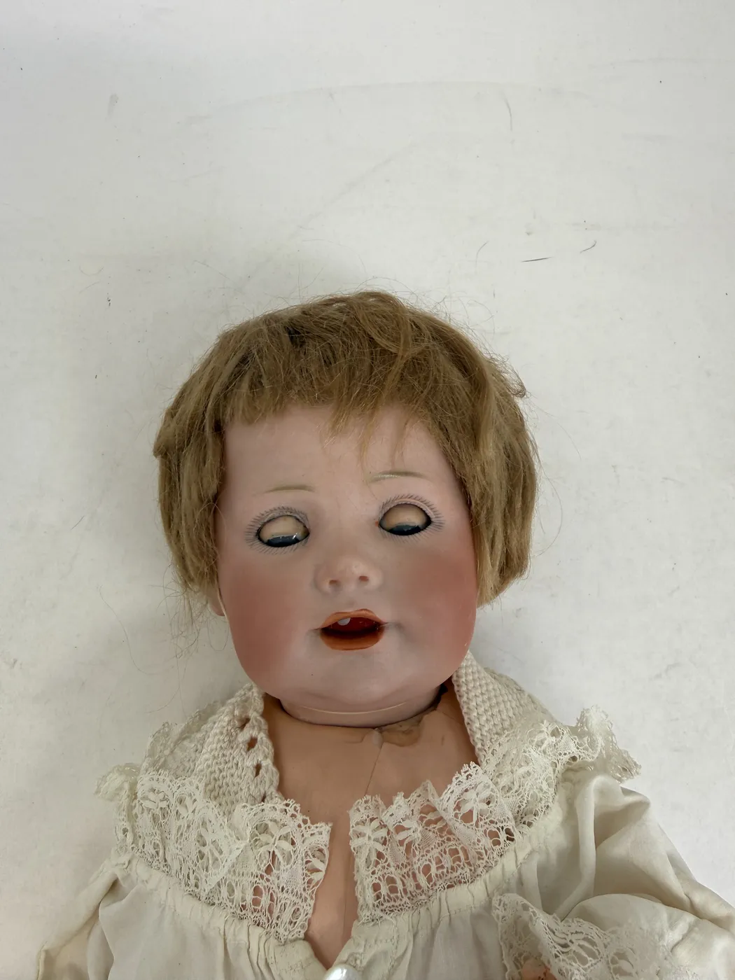 A front-on close-up of the doll's head with the sleep eyes closed, photographed from slightly above with the doll laid on its back. The closed eyelids show the fine painted lower-lash detail crisply, the eye-paint a soft greyish-blue along the lash line. The chubby cheeks are flushed pinker than the surrounding cheek paint, the open mouth shows the two upper teeth and a glimpse of orange tongue, and the mohair wig falls naturally on either side of the temples. The lace-trimmed collar of the christening gown is visible at the bottom of the frame.