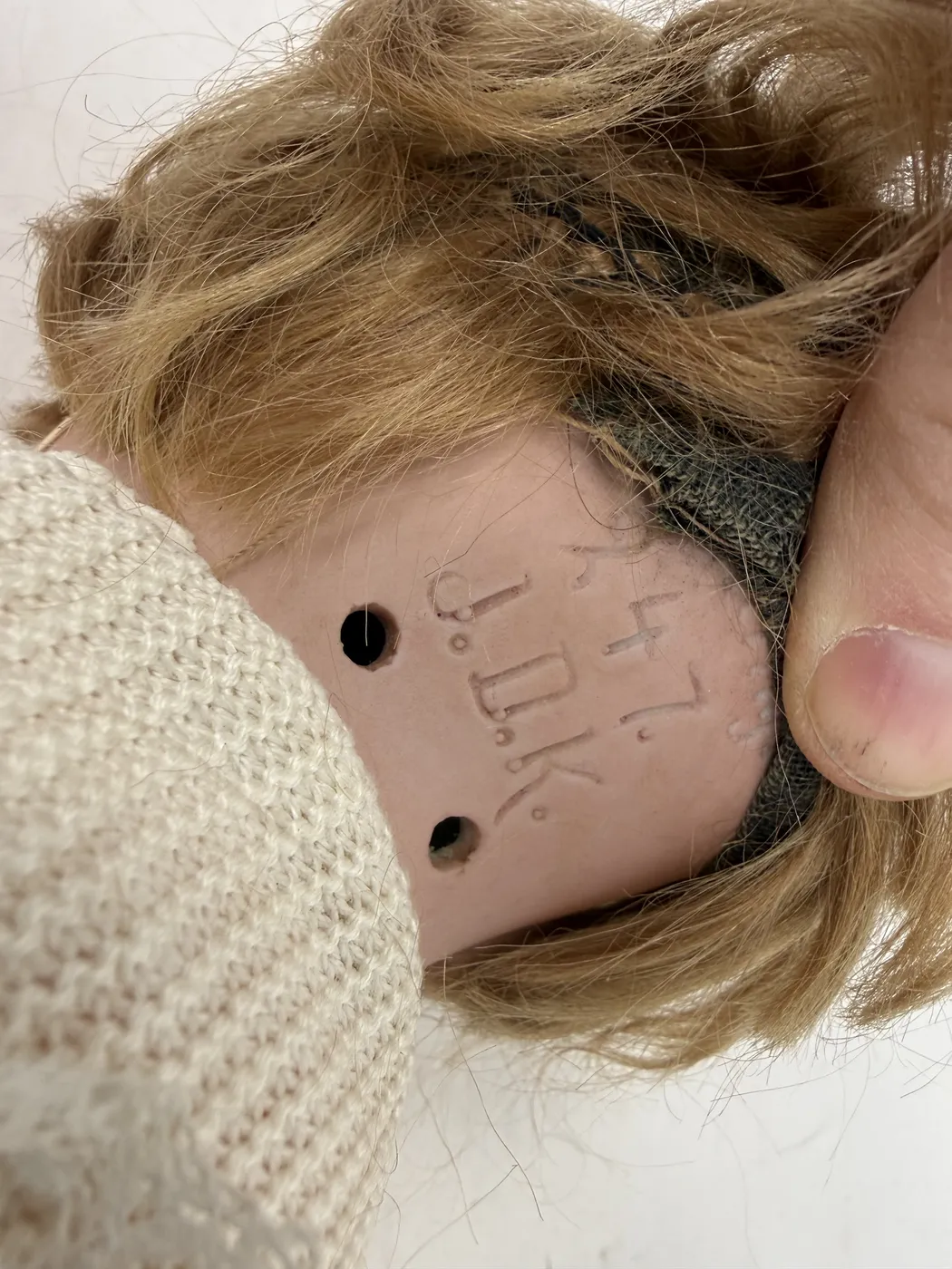 A close-up of the back of the doll's bisque socket head, with the wig and pate gauze pulled aside by a thumb to expose the unglazed bisque underneath. Three lines of incised marking are visible. The top line reads as the numerals 2 4 7, modeled in the slightly serifed continuous-curve font Kestner used for mold numbers in the 1910s. The bottom line reads J . D . K . with periods between each capital. Two round dark stringing holes are visible flanking the marks, the standard location for the elastic stringing that holds the head, body, and limbs in tension. The bisque itself shows the warm pinkish-flesh tone typical of high-grade Kestner character bisque.