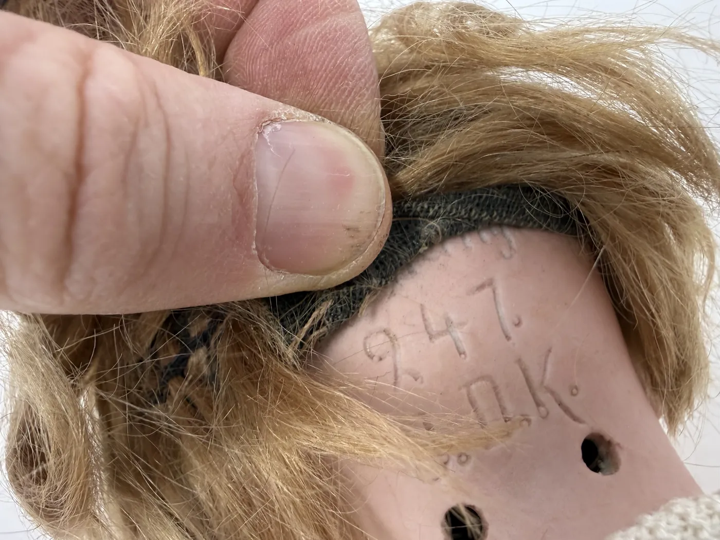 A third mark close-up, this one a more frontal angle on the same back-of-head incised marking, with the wig pulled back from the right side and a thumb at the upper right edge. The same '247' and 'J.D.K.' marks are visible, with a small additional numeral above the 247 that reads approximately as a 9. The two stringing holes are again clearly visible.