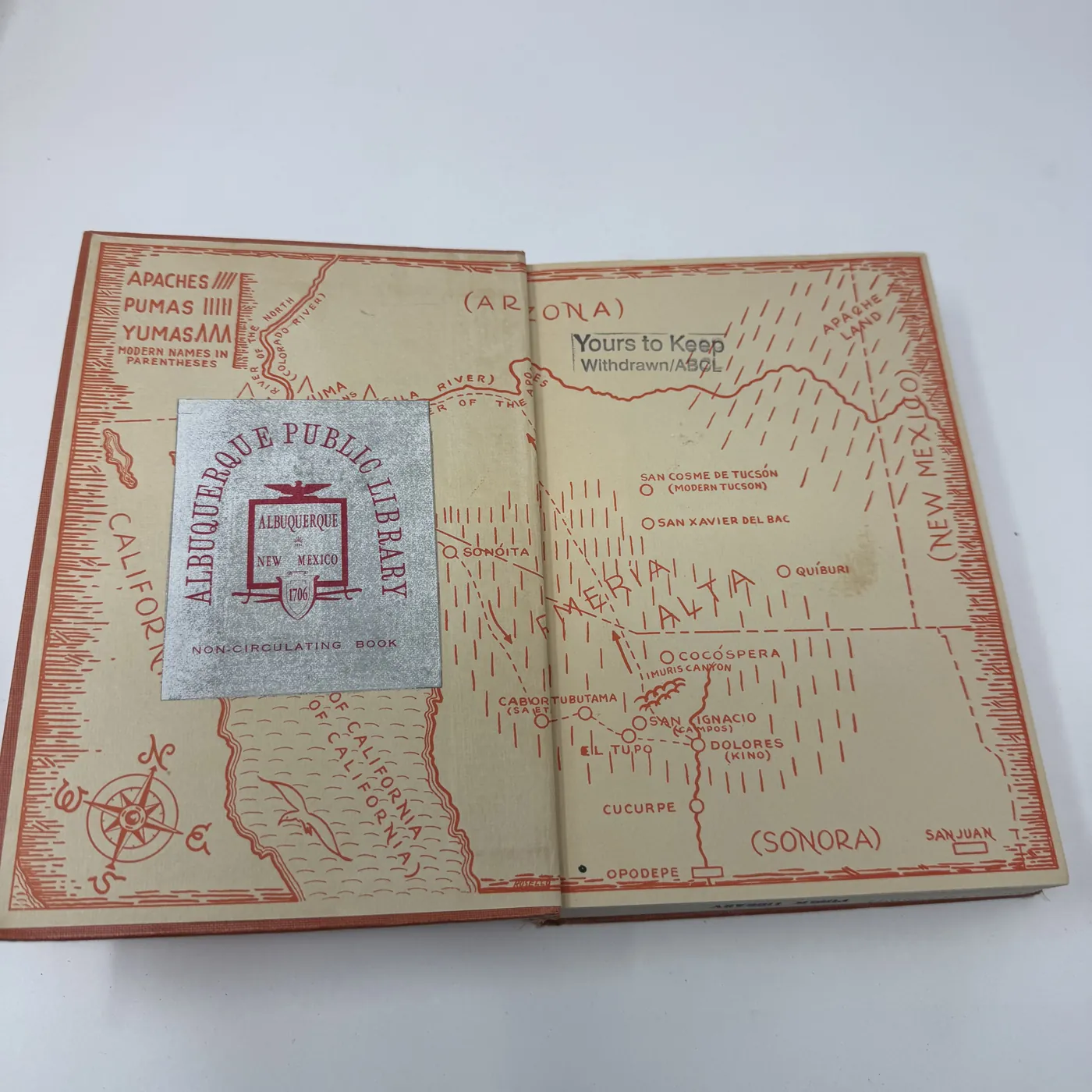 The inside front cover of The Devil's Highway, showing on the left a salmon-ground decorative endpaper map of the Pimería Alta region. Hand-drawn black-line cartography depicts: the Gila River across the top, San Cosme de Tucsón / Modern Tucson and San Xavier del Bac labeled in the upper section, a row of mountains along the New Mexico border on the right; California and the Sea of California labeled across the lower left; Sonora labeled along the bottom; named Spanish mission stations including Sonoita, Caborca, Tubatama, Tubutama, Sáric, San Ignacio, Cocospera (Mision Catedral noted), Magdalena, San Pedro, San Xavier del Bac, Dolores, Cucurpe, Opodepe, San Juan; the tribal labels 'APACHES', 'PUMAS', and 'YUMAS' set in display capitals across the upper portion with the parenthetical 'MODERN NAMES IN PARENTHESES' explanatory legend, and 'ALBUQUERQUE PUBLIC LIBRARY' stamped in a rectangular block at the upper left corner. To the right is the front pastedown, also salmon-pink, plain except for an upside-down 'ALBUQUERQUE PUBLIC LIBRARY' stamp at the lower edge.