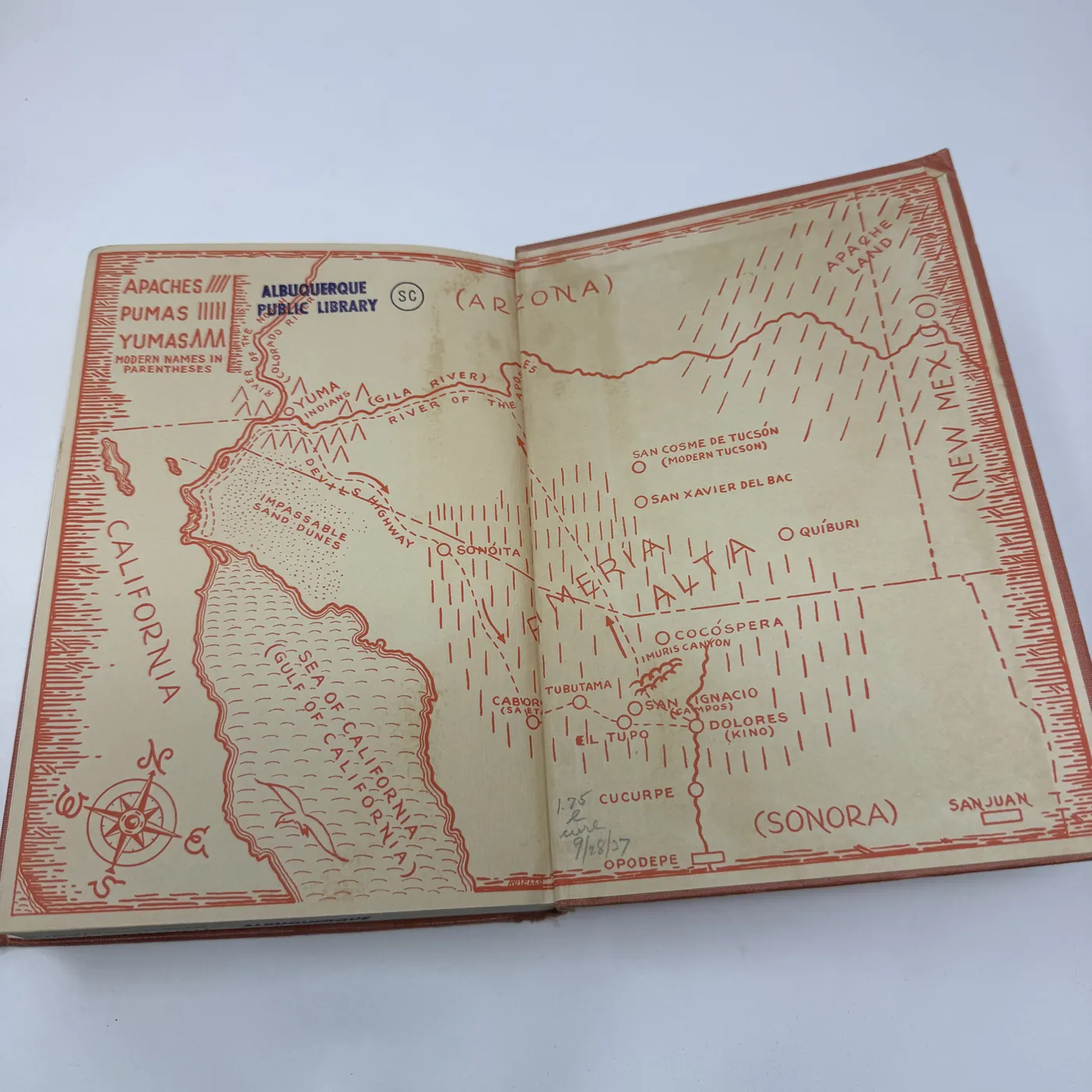 The inside back cover of The Devil's Highway, showing a second salmon-ground decorative cartographic endpaper. The map depicts the Pimería Alta from a slightly different vantage point than the front endpaper: the Gila River runs across the top, a row of black-stippled mountain ranges runs down the right side framing the territory, Sonora and the Sea of California are labeled across the lower portion, and named Spanish mission and town stations including Caborca, San Xavier del Bac, Cocóspera, Magdalena, Dolores, San Pedro, San Ignacio, and Cucurpe are positioned across the territory. A black-line compass rose with a fleur-de-lis north arrow occupies the center-right. The right margin shows the rear-endpaper pastedown, plain salmon, with no markings.