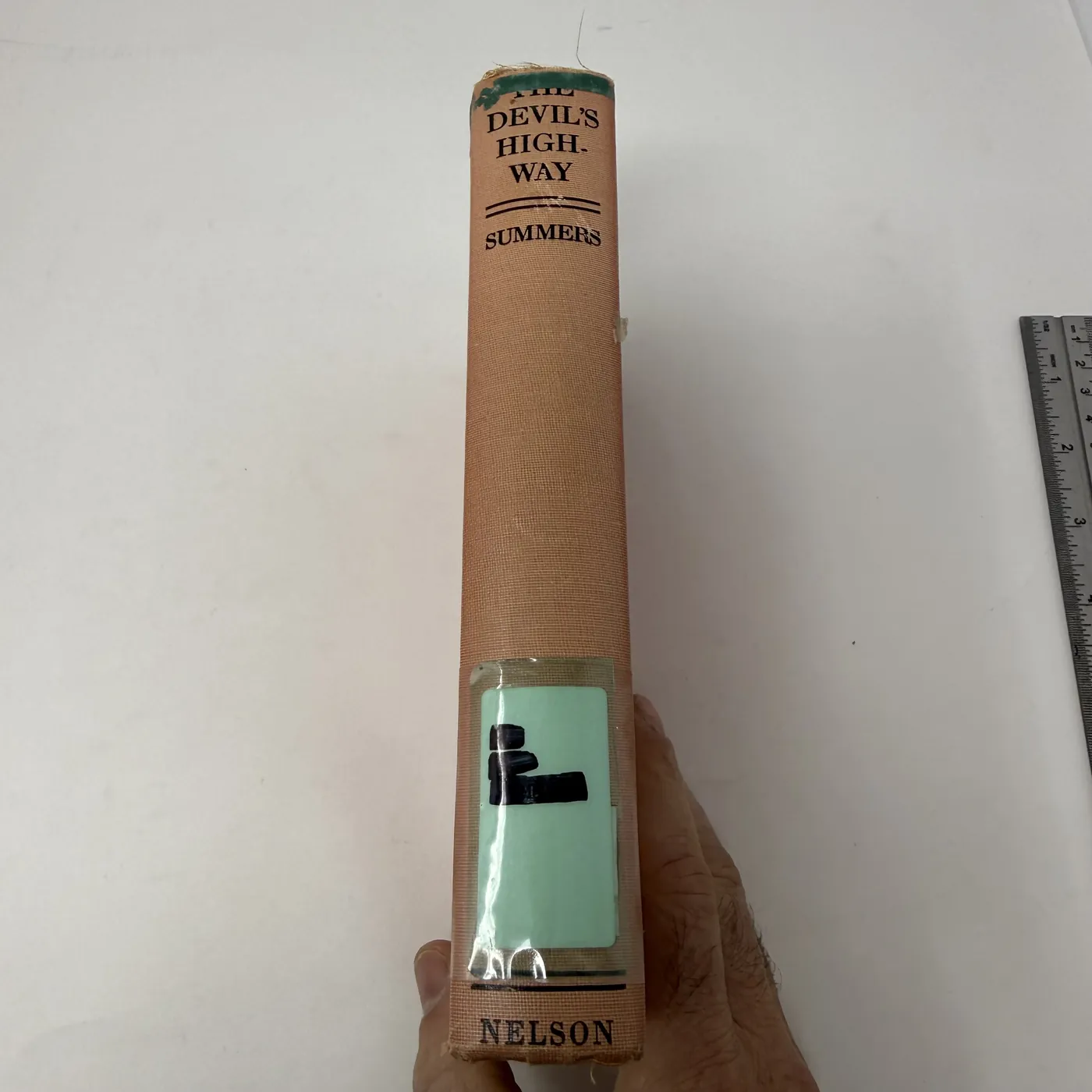 The spine of The Devil's Highway photographed in close detail. The deep terracotta-red cloth runs the length of the spine. At the top, in a small black-stenciled rectangular block: 'THE / DEVIL'S HIGH- / WAY' over a thin horizontal rule then 'SUMMERS'. The body of the spine is plain red cloth, faded near the head and foot from shelf wear. At the foot of the spine in a small black block: 'NELSON' (for the publisher Thomas Nelson and Sons). About one-third up from the foot of the spine, a clear plastic-laminated rectangular Albuquerque Public Library call-number label is affixed, with the visible call number partly obscured by adhesive yellowing but the library label is unmistakably the Albuquerque institutional format.
