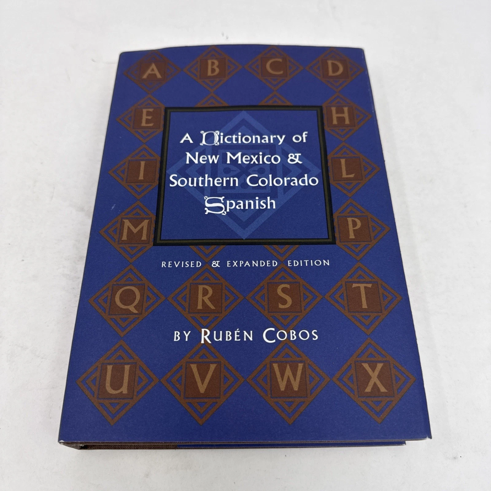 The blue dust jacket of A Dictionary of New Mexico and Southern Colorado Spanish by Rubén Cobos, with stylized alphabet motif on the cover, Revised & Expanded Edition.