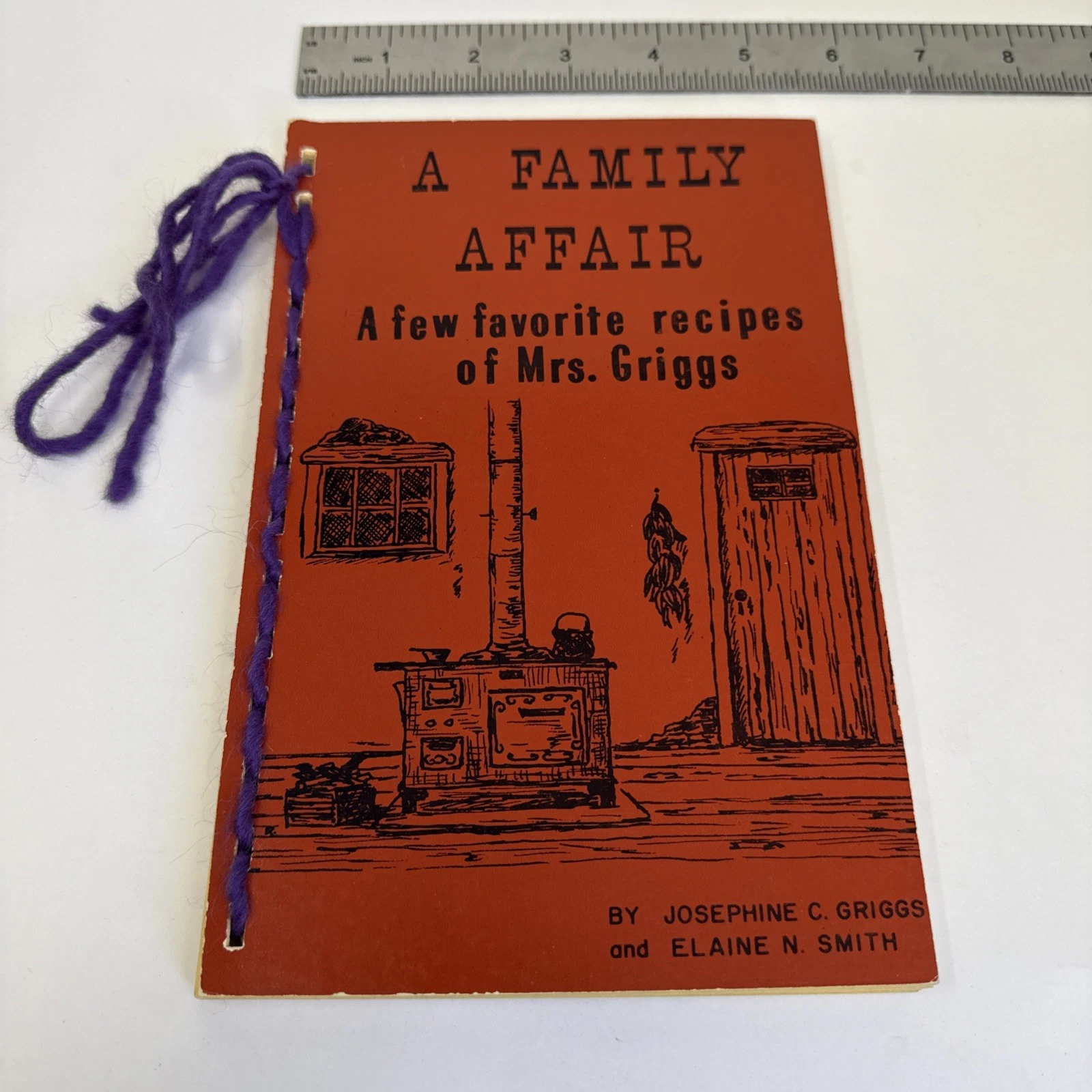 A small orange paperback cookbook with purple yarn binding titled 'A Family Affair: A Few Favorite Recipes of Mrs. Griggs' by Josephine C. Griggs and Elaine N. Smith, with a pen-and-ink illustration of a wood-burning stove with chiles drying overhead.