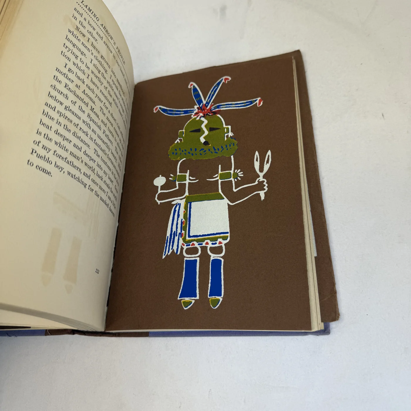 One of James Paytiamo's color drawings: a dancer in a green mask-like face with feathered headdress, white kilt and blue moccasins, reproduced on a brown ground.