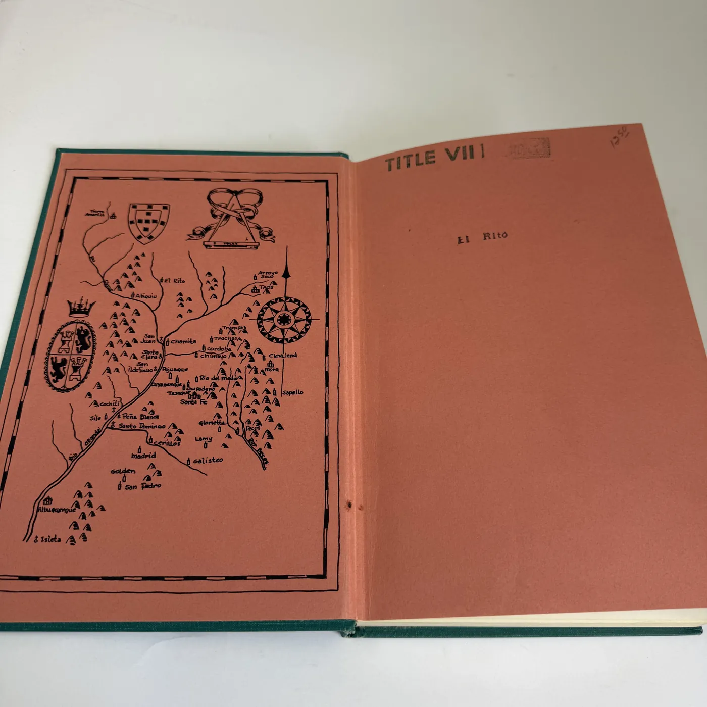 The inside front cover of the book showing a salmon-pink ground decorative endpaper map of the Upper Rio Grande valley with hand-drawn black-line landmarks: a compass rose with cardinal points, two coat-of-arms cartouches (Spanish heraldry of Castile and León), the river running south through clusters of cottonwood and piñon hills, and the named villages including Albuquerque, San Pedro, Madrid, Galisteo, Santa Fe, Tesuque, San Ildefonso, Chimayó, Las Trampas, Truchas, Santa Cruz, El Rito, and the Cordova / San Juan / Ohkay Owingeh area. To the right, a plain salmon-pink front pastedown with the typed words 'TITLE VII' at the top corner and 'El Rito' written in pencil in the center as a reader's chapter marker.