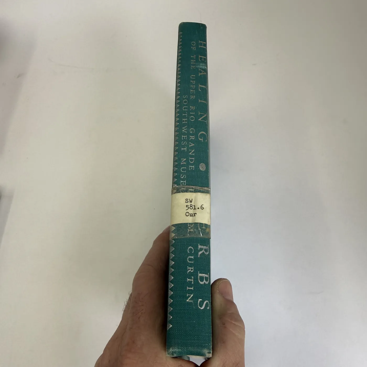 The spine of the book photographed in close detail, showing the dark green book-cloth with the title 'HEALING OF THE UPPER RIO GRANDE / SOUTHWEST MUSEUM' stamped in silver running vertically along the spine and 'HERBS / L S M / CURTIN' stamped in the lower portion. A small white Albuquerque Public Library call-number label is affixed near the foot of the spine reading: 'SW / 581.6 / Cur'.