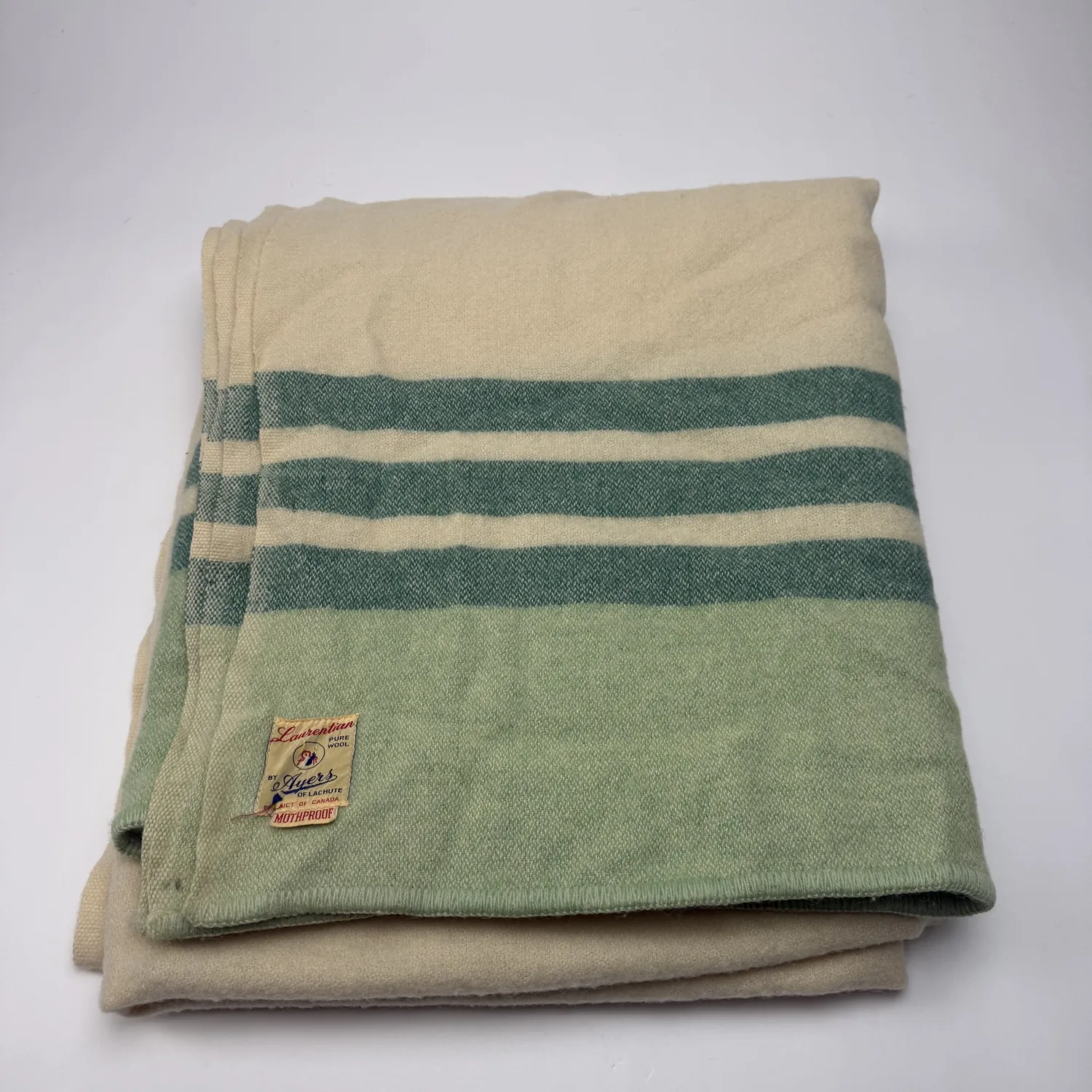Folded cream and sage-green Laurentian Pure Wool blanket by Ayers of Lachute, with twin forest-green stripes and the original yellowed woven label (red Mothproof banner, Product of Canada) visible on the green section.