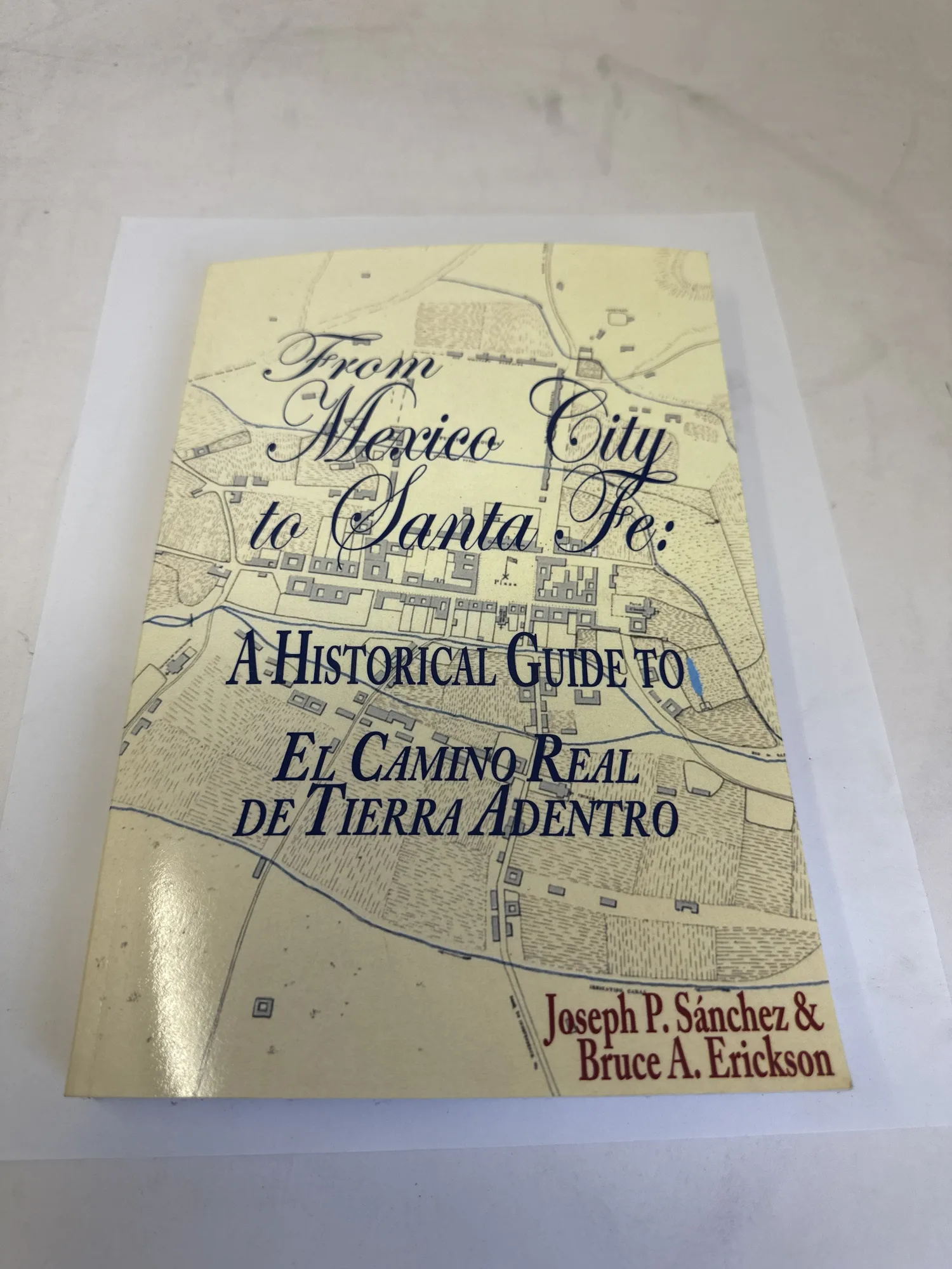 Cover of From Mexico City to Santa Fe: A Historical Guide to El Camino Real de Tierra Adentro by Joseph P. Sánchez and Bruce A. Erickson, showing a historical colonial-era map of a Spanish settlement as the background with ornate calligraphic title typography. Published by Rio Grande Books. Photographed at the New Mexico Literacy Project sorting desk in Albuquerque.