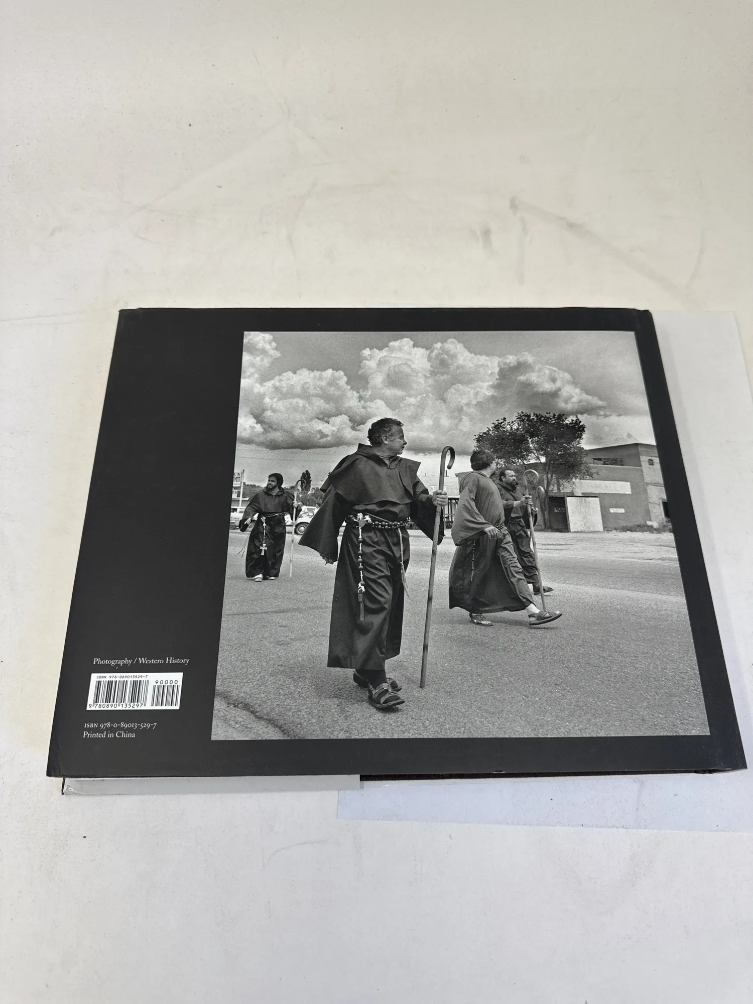 Back cover of In Search of Domínguez and Escalante showing black-and-white photograph of two Franciscan friars walking through a pueblo village with dramatic cloud-filled sky above. Photography / Western History category. Photographed at the New Mexico Literacy Project sorting desk in Albuquerque.