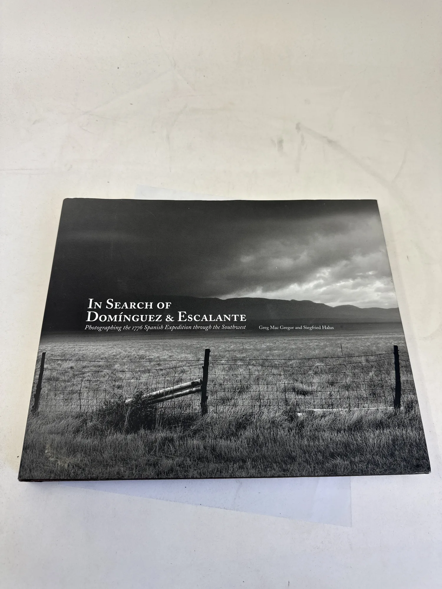 Front cover of In Search of Domínguez and Escalante by Greg Mac Gregor and Siegfried Halus, Museum of New Mexico Press 2011 — dramatic black-and-white landscape photograph of Southwest desert with wooden crosses in foreground and storm clouds above. Photographed at the New Mexico Literacy Project sorting desk in Albuquerque.