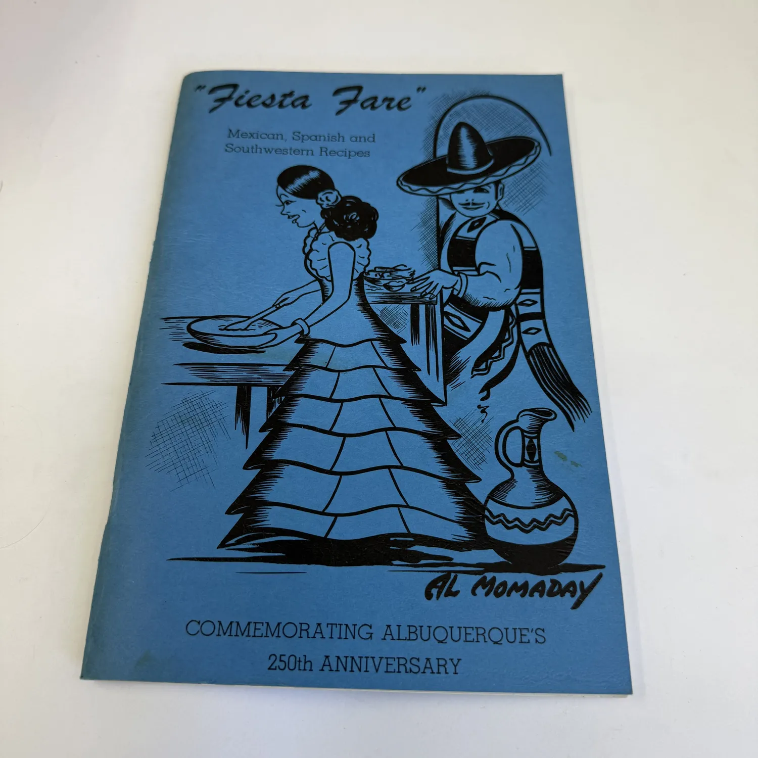 A 1956 Fiesta Fare cookbook for the Albuquerque 250th anniversary celebration, with cover art credited to Al Momaday, lying on a wooden surface.