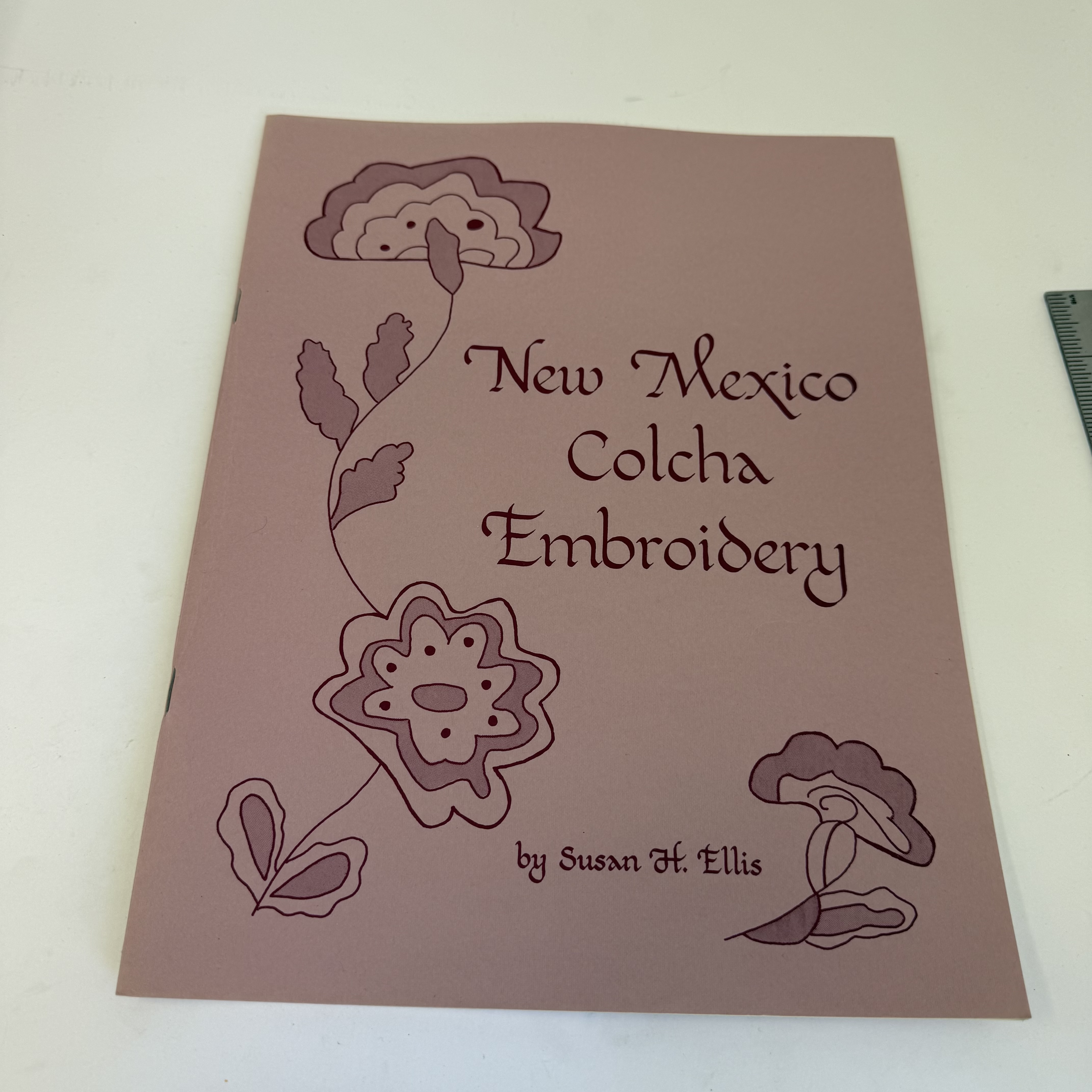 A pink-cover specialty book 'New Mexico Colcha Embroidery' by Susan H. Ellis, a regional handbook on traditional New Mexico Hispanic colcha embroidery, donated to NMLP after a library asked the donor to sort first.