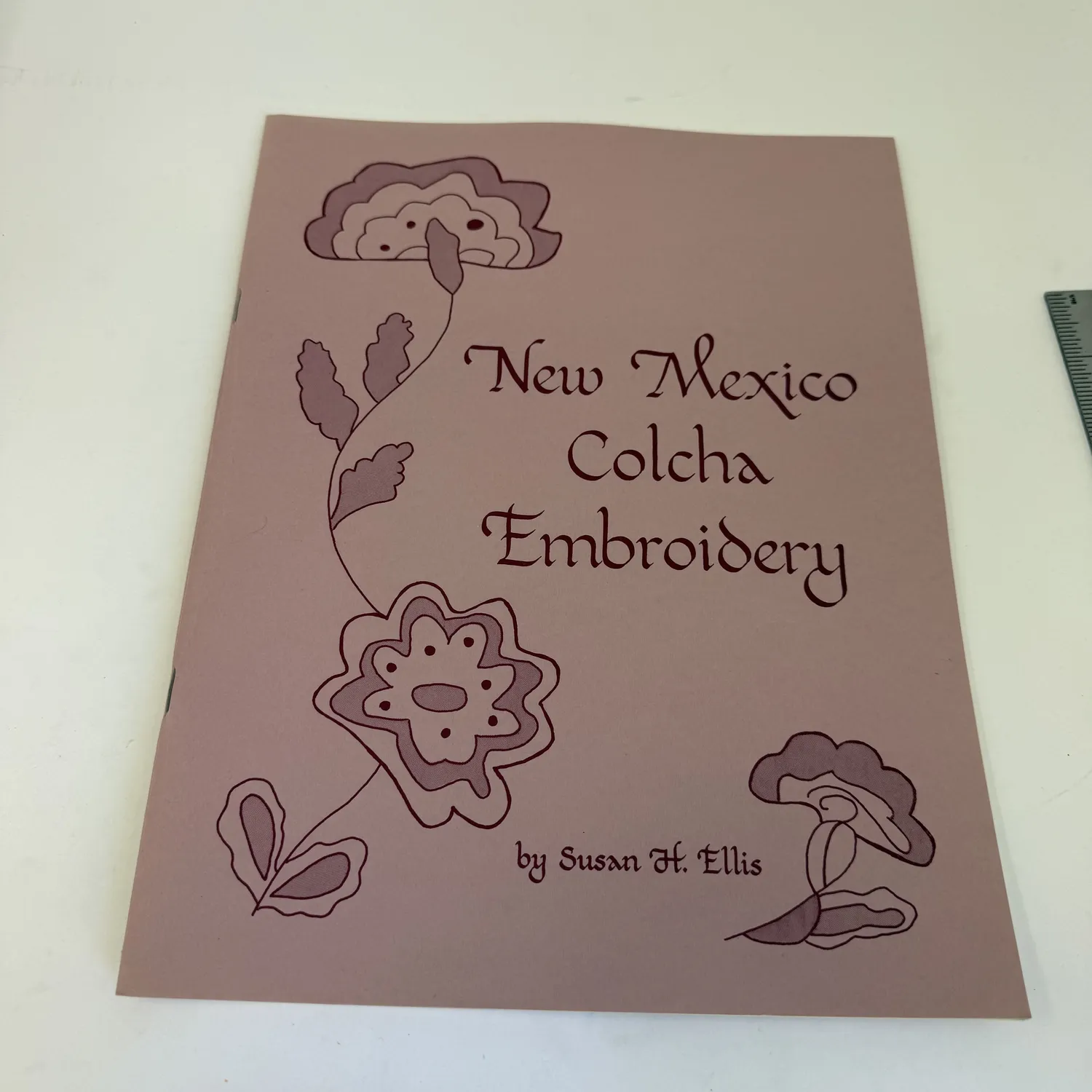 A copy of New Mexico Colcha Embroidery by Susan H. Ellis lying on a wooden surface, a saddle-stitched paperback technical handbook on the traditional NM Hispanic embroidery technique.