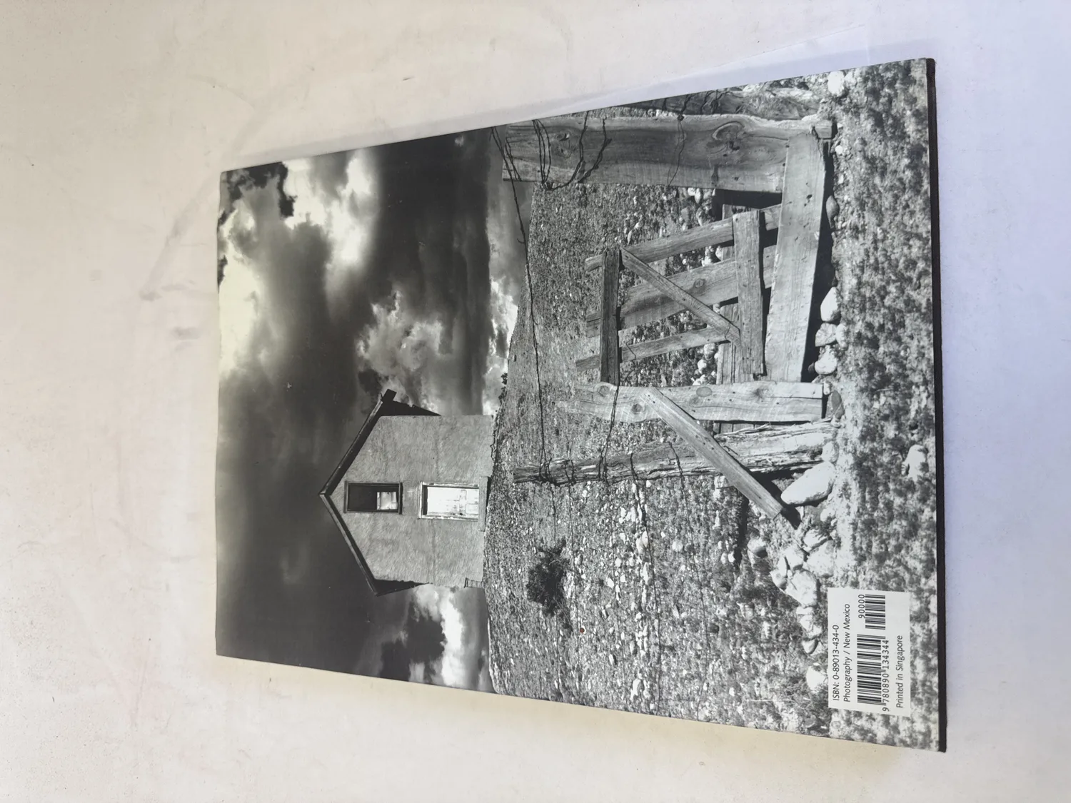 Back cover of Ernest Knee in New Mexico showing a black-and-white photograph of a rural New Mexico ranch structure with wooden pole fence and dramatic cumulus clouds, barcode with ISBN 0-89013-434-0, printed in Singapore. Photographed at the New Mexico Literacy Project sorting desk in Albuquerque.