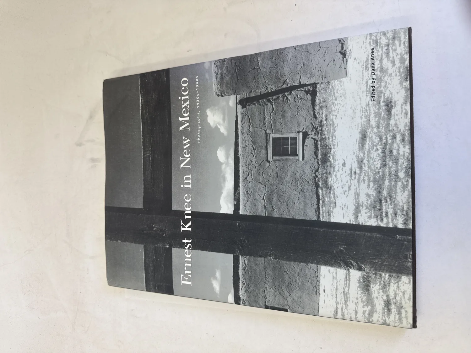 Front cover of Ernest Knee in New Mexico Photographs 1930s-1940s showing a black-and-white gelatin silver print of an adobe building with a single window and dramatic cloud-streaked sky, edited by Dana Knee, Museum of New Mexico Press 2005. Photographed at the New Mexico Literacy Project sorting desk in Albuquerque.
