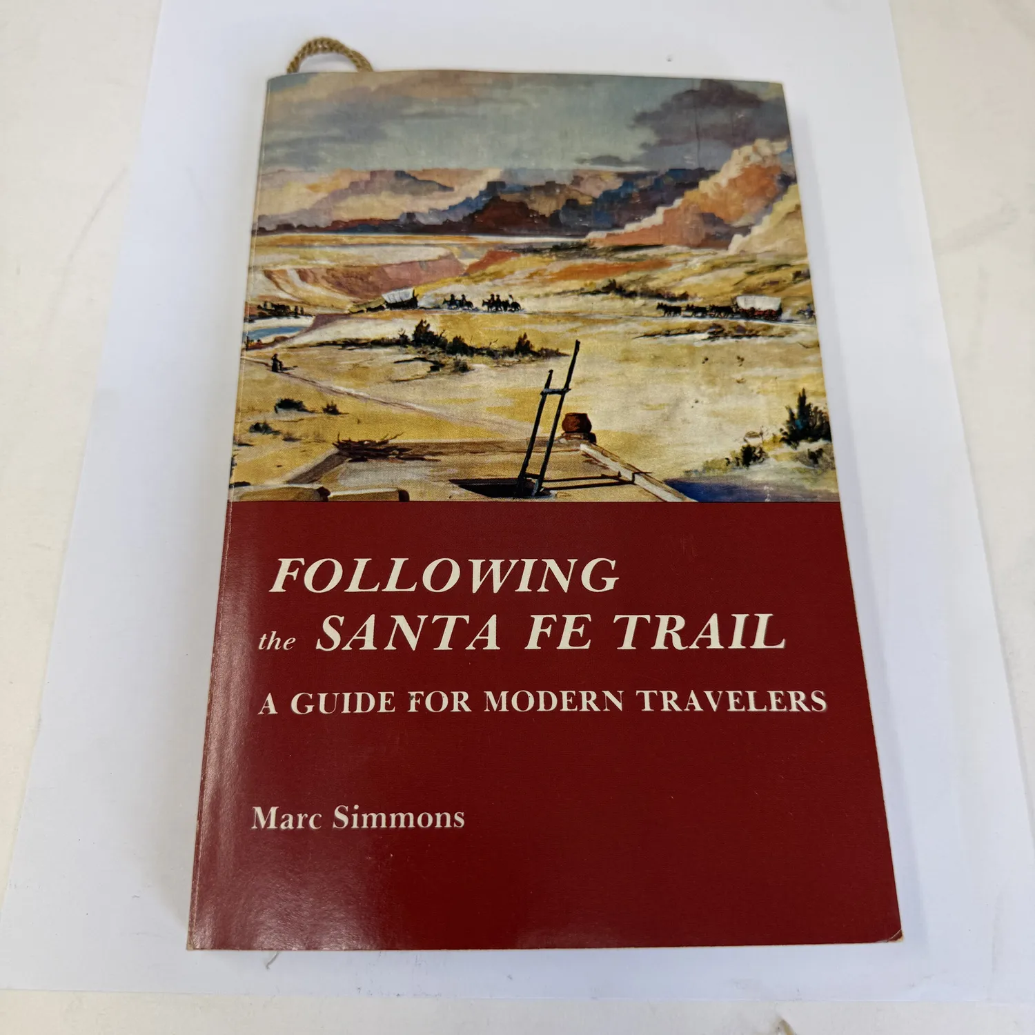 Cover of Following the Santa Fe Trail A Guide for Modern Travelers by Marc Simmons — watercolor illustration of a wagon train crossing the prairie with mesas and mountains in the background, red lower panel with title text. Found in an Albuquerque estate donation to the New Mexico Literacy Project.