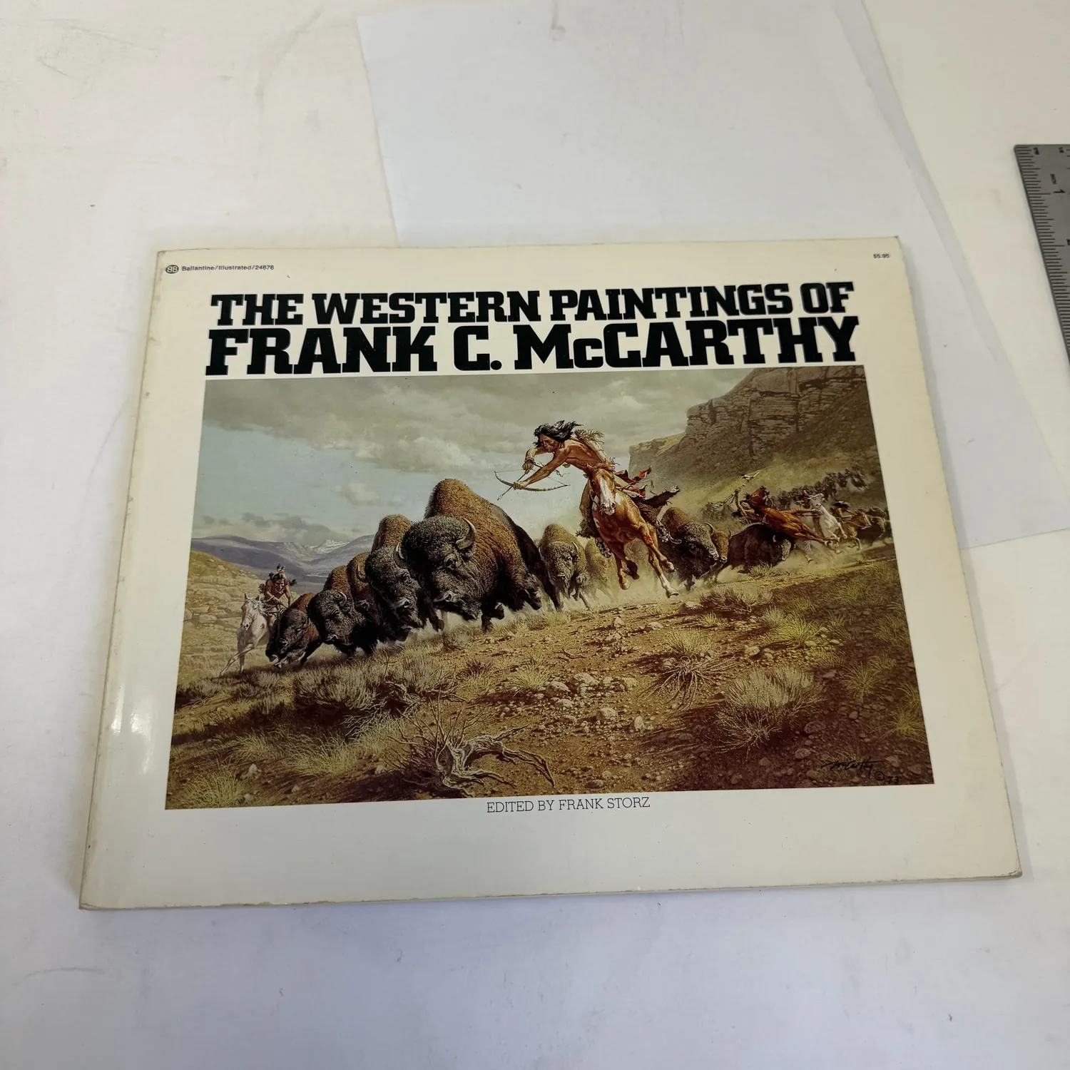 Front cover of The Western Paintings of Frank C. McCarthy, edited by Frank Storz, Ballantine Books. Large-format softcover showing The Sioux Buffalo Run (1973), a dramatic cavalry-and-buffalo action painting in warm earth tones. Title in white and gold lettering. Fourth Printing, January 1976. Photographed at the New Mexico Literacy Project sorting desk in Albuquerque.