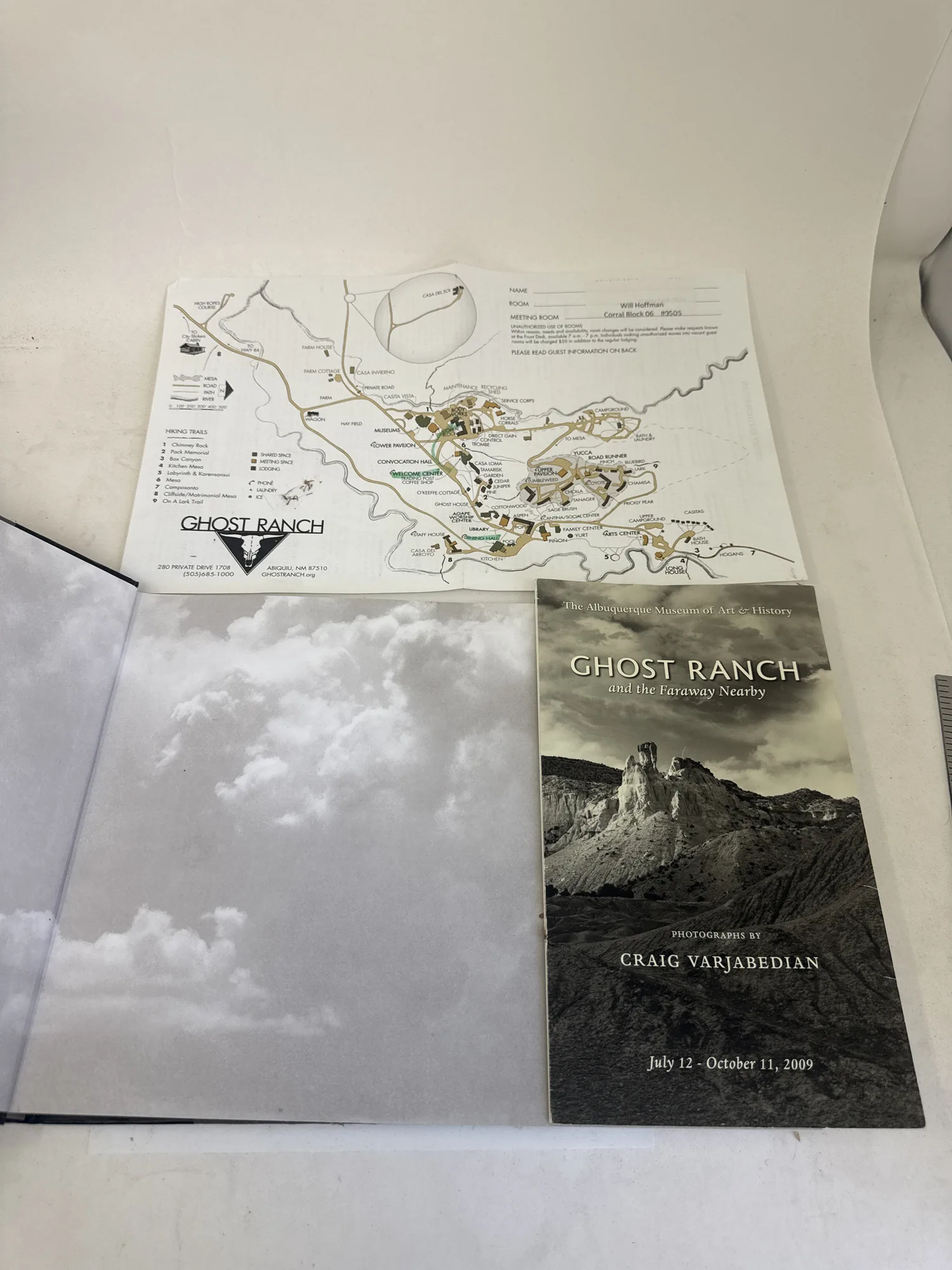 Folded Ghost Ranch trail and property map insert alongside the back cover of Ghost Ranch and the Faraway Nearby, showing exhibition dates July 12 through October 11 2009 at the Albuquerque Museum of Art and History. Photographed at the New Mexico Literacy Project sorting desk in Albuquerque.
