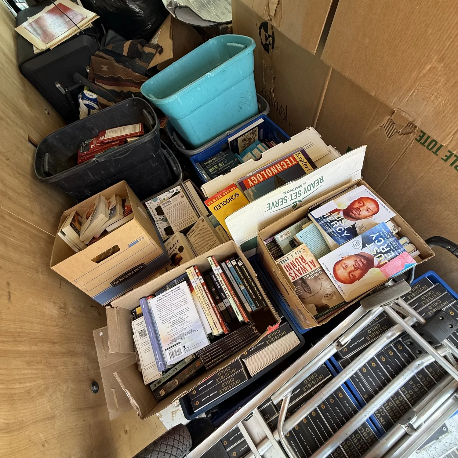 A second angle of the loaded van showing open boxes of Gilbert Sena classroom books — the Just Mercy young-adult adaptation, Always Running, Schooled, a Ready-Set-Serve culinary box, and rows of Invisible Child hardcovers — beside totes and surplus property cleared from the school.
