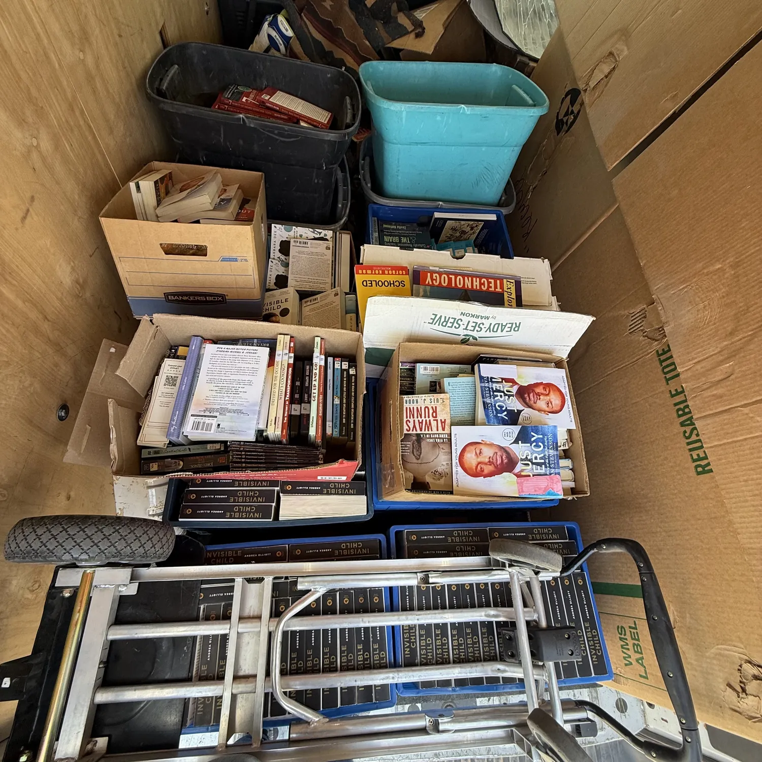 The bed of the NMLP cargo van loaded with banker boxes and totes of donated classroom books from Gilbert L. Sena Charter High School — visible class sets and titles include Invisible Child, the Just Mercy young-adult adaptation, Always Running, Schooled, and a Technology reference text, with a hand truck staged in front.