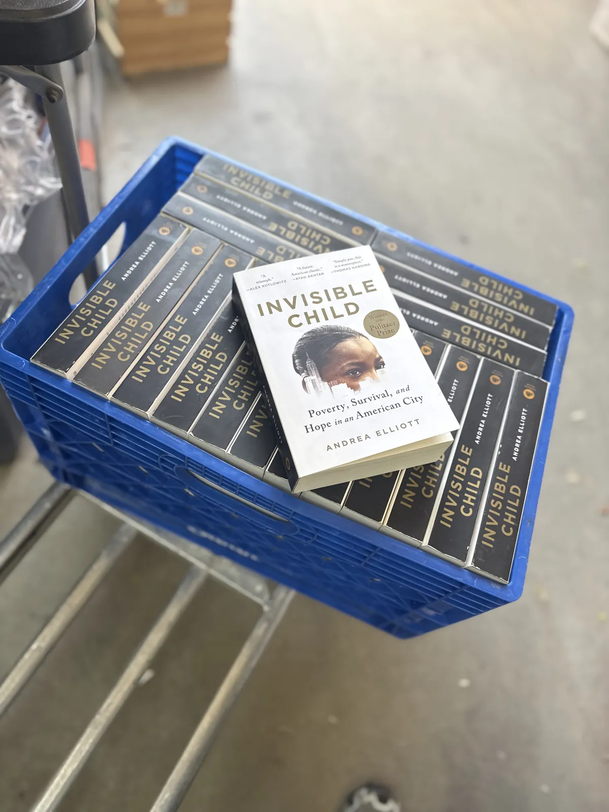 A blue crate stacked with a full class set of Invisible Child by Andrea Elliott on a warehouse cart, with one copy stood up face-out showing the Pulitzer Prize seal — the set routed from NMLP to the Read to Me! ABQ Network book room for Albuquerque teacher book clubs and students.