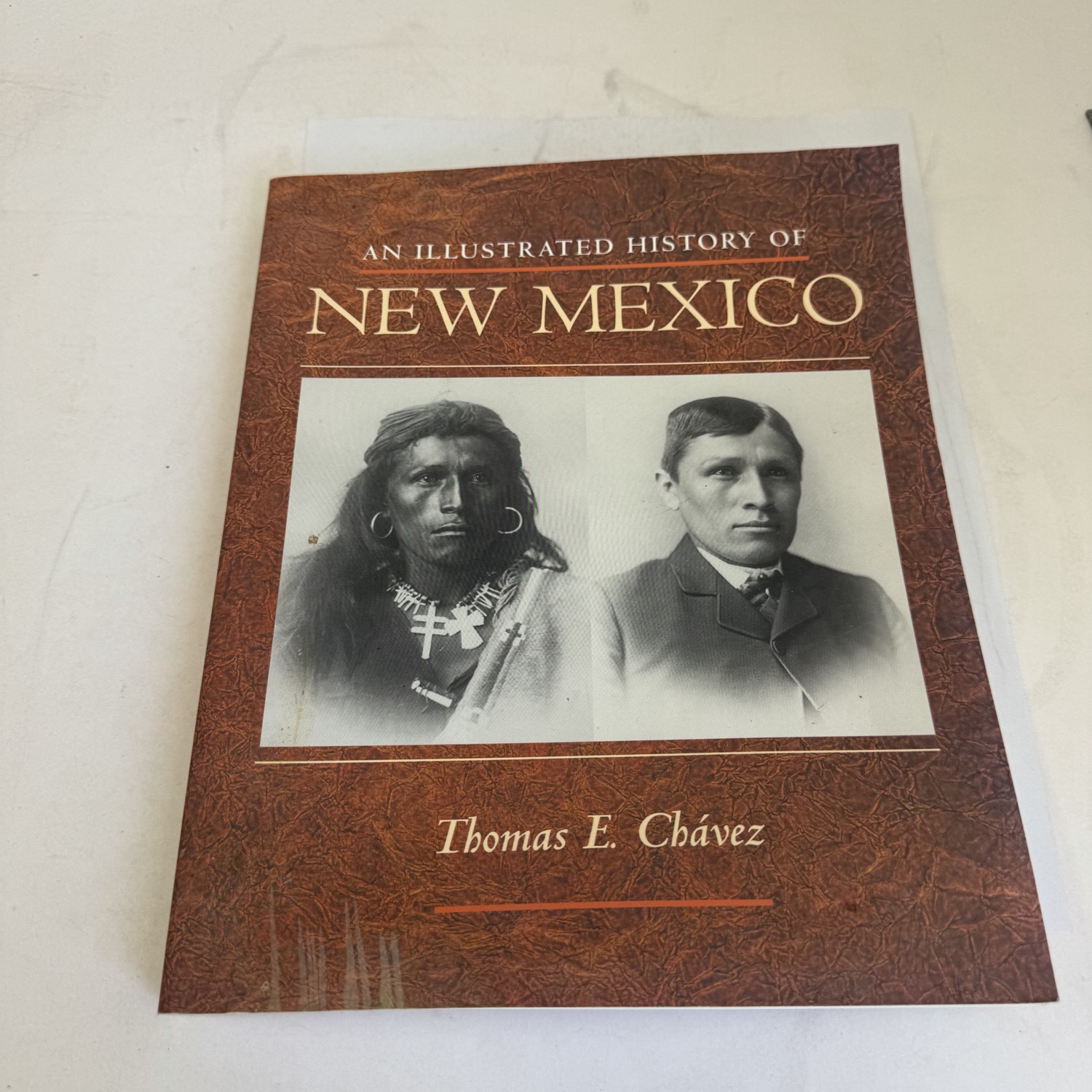 Front cover of An Illustrated History of New Mexico by Thomas E. Chávez — brown leather-textured UNM Press paperback with two historical black-and-white portraits of Tony Todaro (Navajo) photographed at the Carlisle Indian School ca. 1884 and ca. 1887, gold lettering, cover design by Robyn Mundy