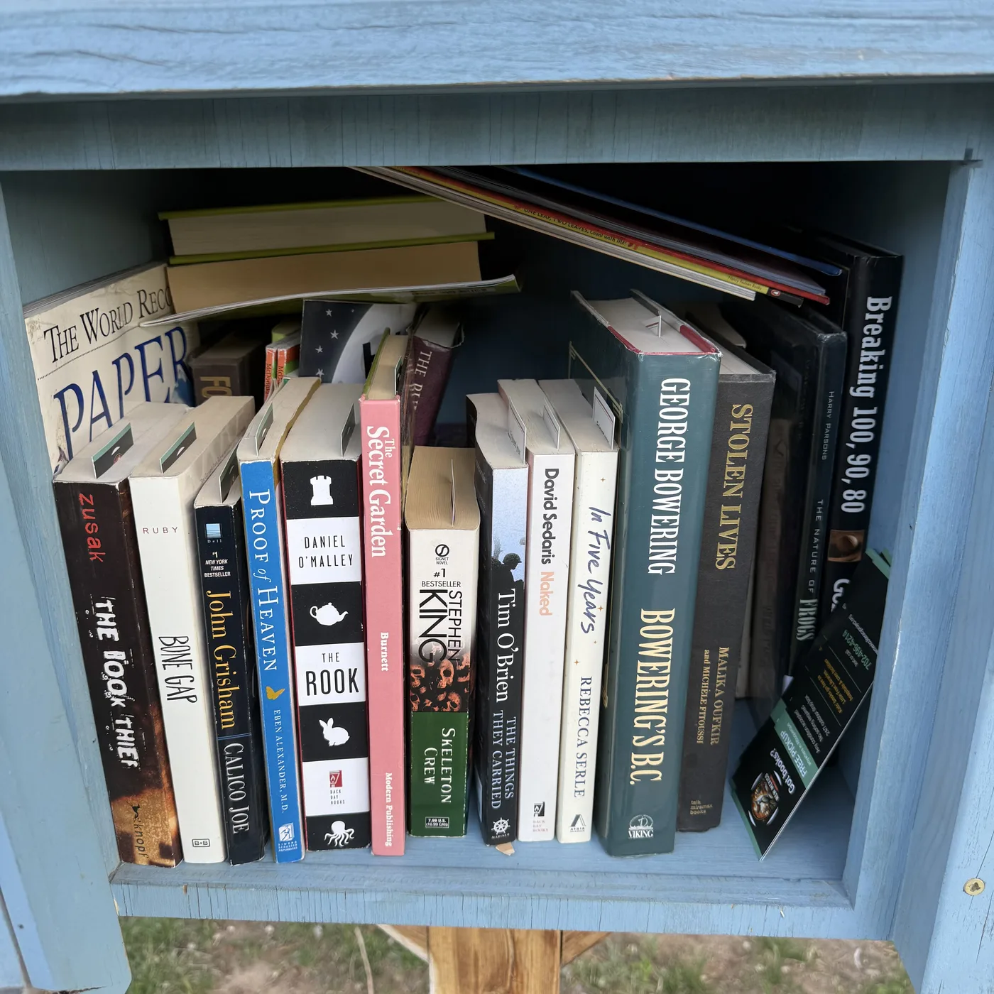 A blue Little Free Library shown from the front, deep main shelf packed with adult fiction and nonfiction restocked from NMLP intake. Visible spines (left to right): Markus Zusak 'The Book Thief' (Knopf), Laura Ruby 'Bone Gap' (Balzer + Bray), John Grisham 'Calico Joe' (Dell #1 NYT Bestseller), Eben Alexander MD 'Proof of Heaven' (Simon & Schuster), Daniel O'Malley 'The Rook' (Back Bay Books), Frances Hodgson Burnett 'The Secret Garden' (Modern Publishing pink edition), Stephen King 'Skeleton Crew' (Signet mass market), Tim O'Brien 'The Things They Carried' (Mariner), David Sedaris 'Naked' (Back Bay Books), Rebecca Serle 'In Five Years' (Atria), George Bowering 'Bowering's B.C.' (Viking hardcover green), 'Stolen Lives' by Malika Oufkir and Michèle Fitoussi (Talk Miramax Books hardcover), Harry Parsons 'The Nature of Frogs' hardcover, 'Breaking 100, 90, 80' golf-instruction hardcover. Background top shelf: 'The World Record Paper...' airplane book, food and other reference. Bottom right corner: black NMLP 'Got books? Free pickup' bookmark visible jutting out of the shelf right edge.
