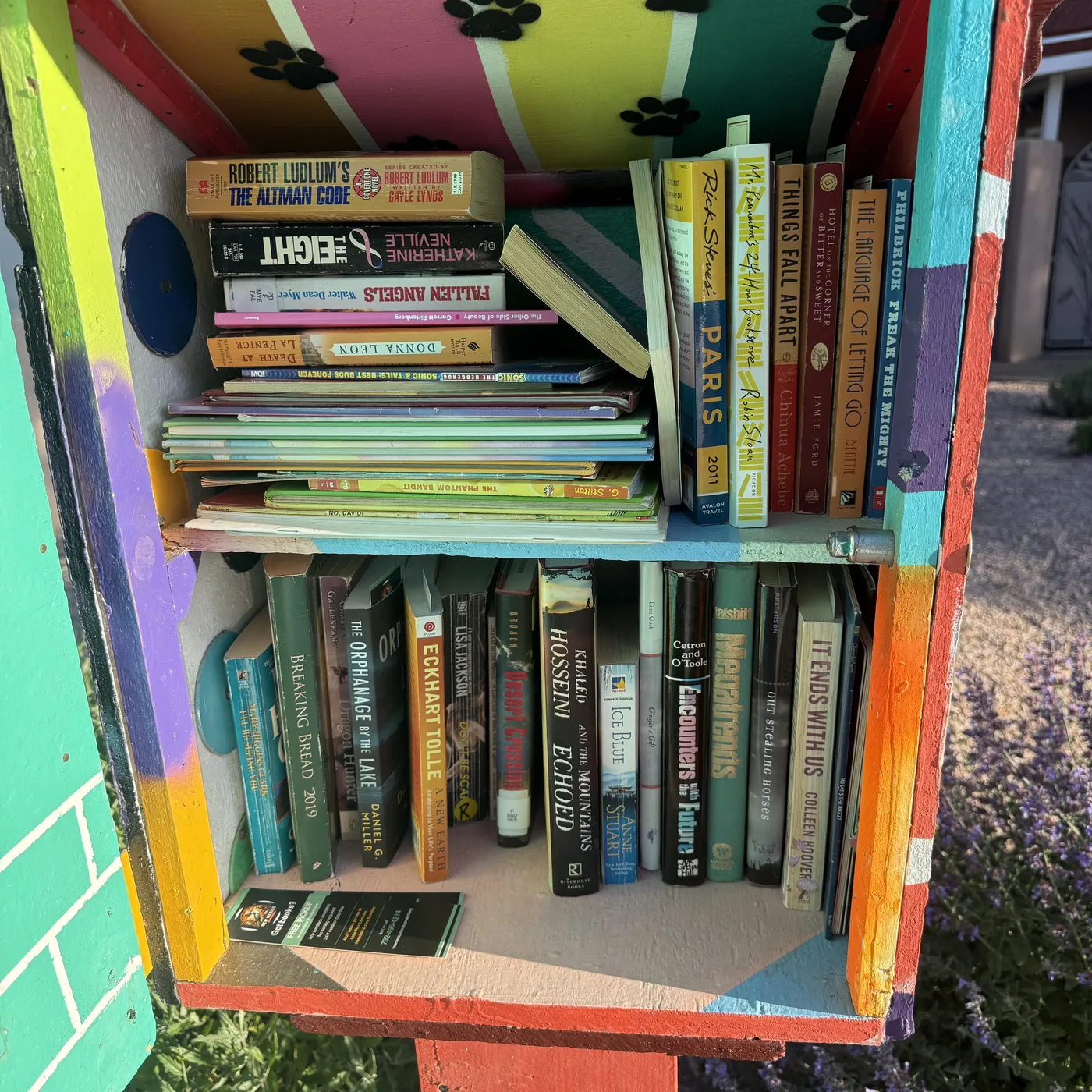 A blue painted Little Free Library with two glass-paneled doors, showing two shelves of books restocked from NMLP intake. Top shelf: Liane Moriarty 'Truly Madly Guilty', David Baldacci 'The Last Mile', vintage Thorndike Comprehensive Desk Dictionary volumes A-K and L-Z (the original beige two-volume reference), 'Top 20 Daredevils', Carrie Stuart Parks 'Fairy Unicorns', Dr. Seuss / Rosetta Stone 'Because a Little Bug Went Ka-CHOO!' standing face out, with backup books behind. Right pane shows Richard Russo 'Fallen' hardcover, Vanderbilt fantasy, Doonesbury Chronicles, more. Bottom shelf: Crandace Camp 'The Bridal Quest', vintage 'Dave Darrin's First Year at Annapolis' (Saalfield), Roget's Thesaurus, Edgar Leoni 'Nostradamus', 'Summer of Champions', Dewey Johnson 'Drag Racing Funny Cars', 'Crimes and Punishment', Maeve Binchy, an NMLP bookmark visible at center showing 'Got books? Free pickup' and the phone number 702-496-4214.